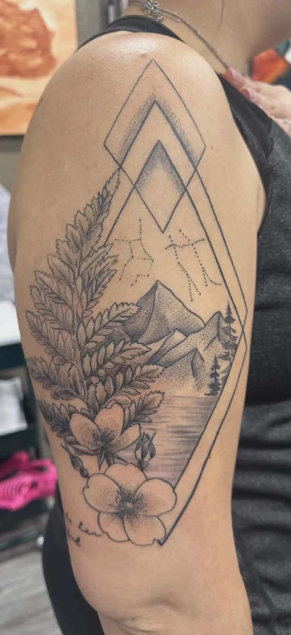Tattoo of mountains, trees, a lake, a large leaf, and a flower with geometric lines and shading on a person's upper arm.