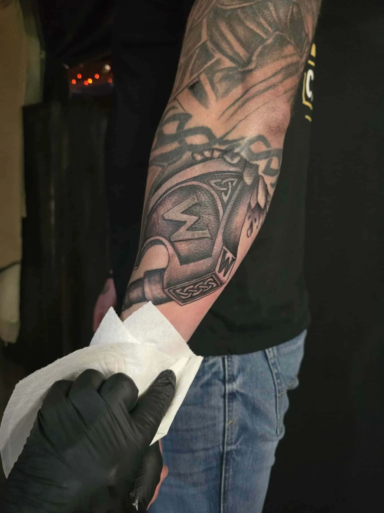 Close-up of a person receiving a tattoo on their forearm, depicting a hammer with a heart and detailed designs, with a gloved hand holding a tissue for cleaning or wiping.