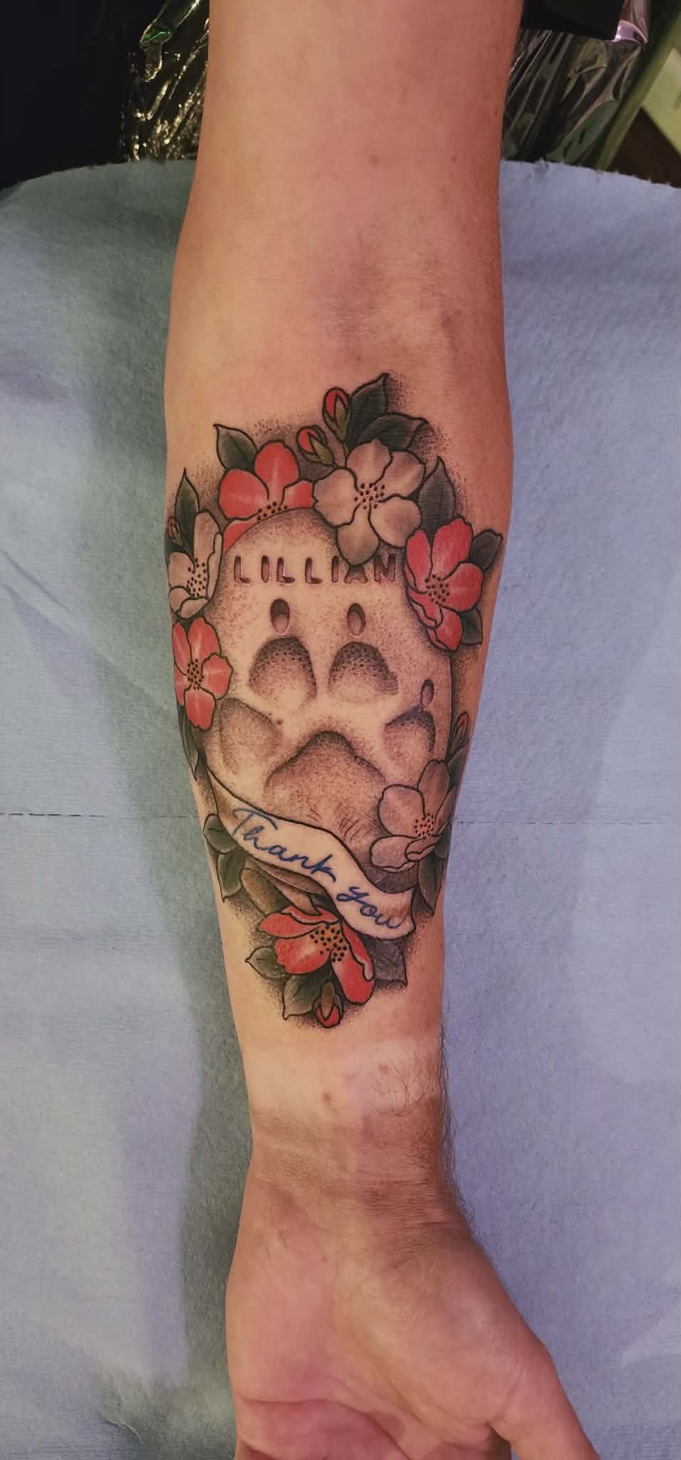 Tattoo of a small dog skull surrounded by pink and white flowers with green leaves, and a ribbon below that says "Thank you" in blue script.