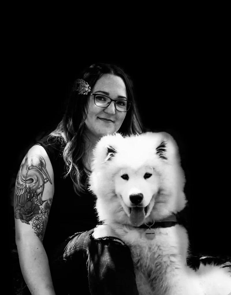 A woman with glasses and tattoos on her arm, smiling with a fluffy white dog, possibly a Samoyed, against a black background.