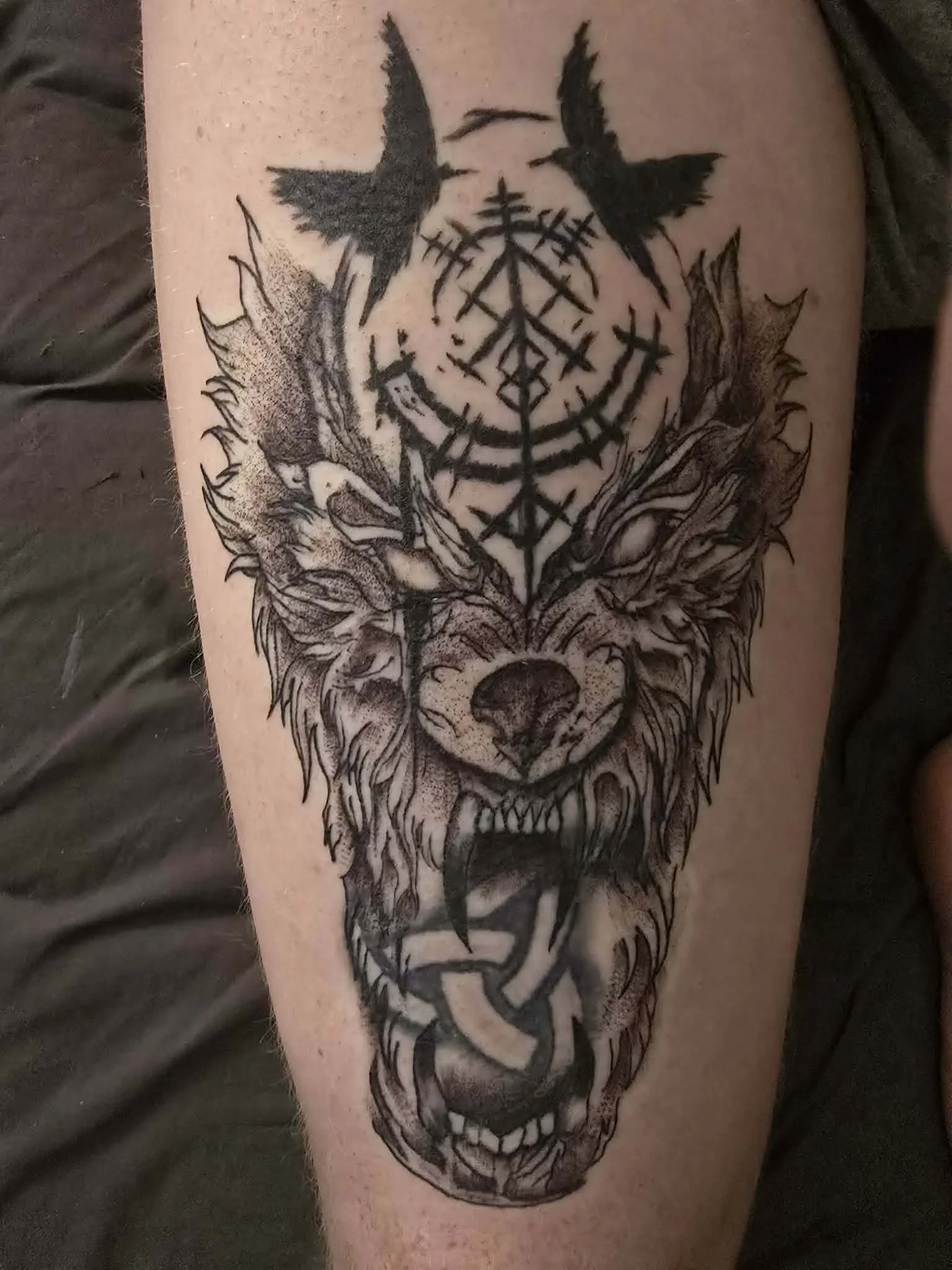 A detailed tattoo of a snarling wolf's head with its mouth open, sharp teeth visible, and eyes intense, with intricate line work and shading. Above the wolf's head, there are two black birds flying and a circle with occult symbols and geometric patterns inside.