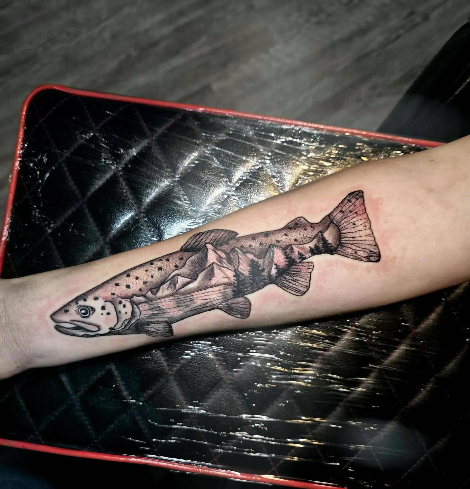 A tattoo of a fish with mountain scenery and trees inside it, on a person's forearm.