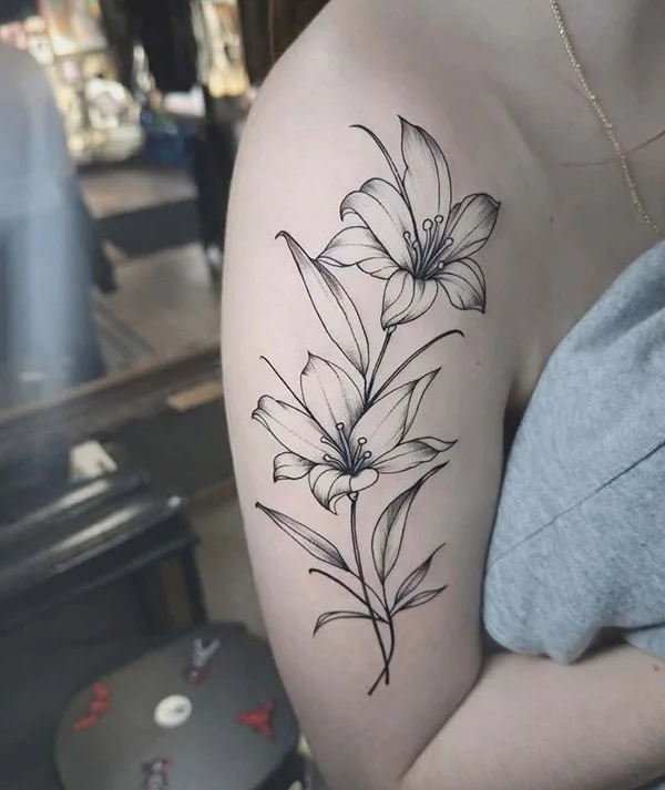 Black and gray outline tattoo of two lilies on a person's upper arm, with detailed petals and leaves.