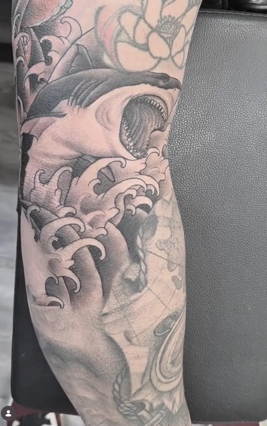 Close-up of a tattoo sleeve featuring a large, roaring shark with waves and ocean elements around it, as well as other floral and map details.