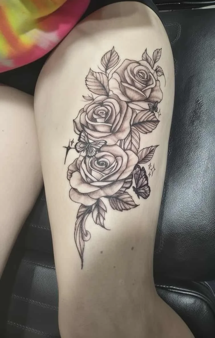 Black and gray tattoo of three roses with leaves and two butterflies on a person's thigh.
