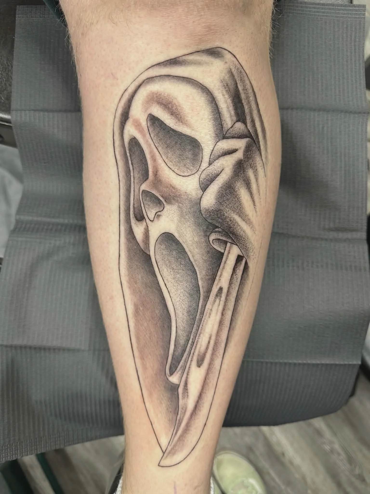Black and gray tattoo of a scream mask on a person's calf, with shading and fine lines.