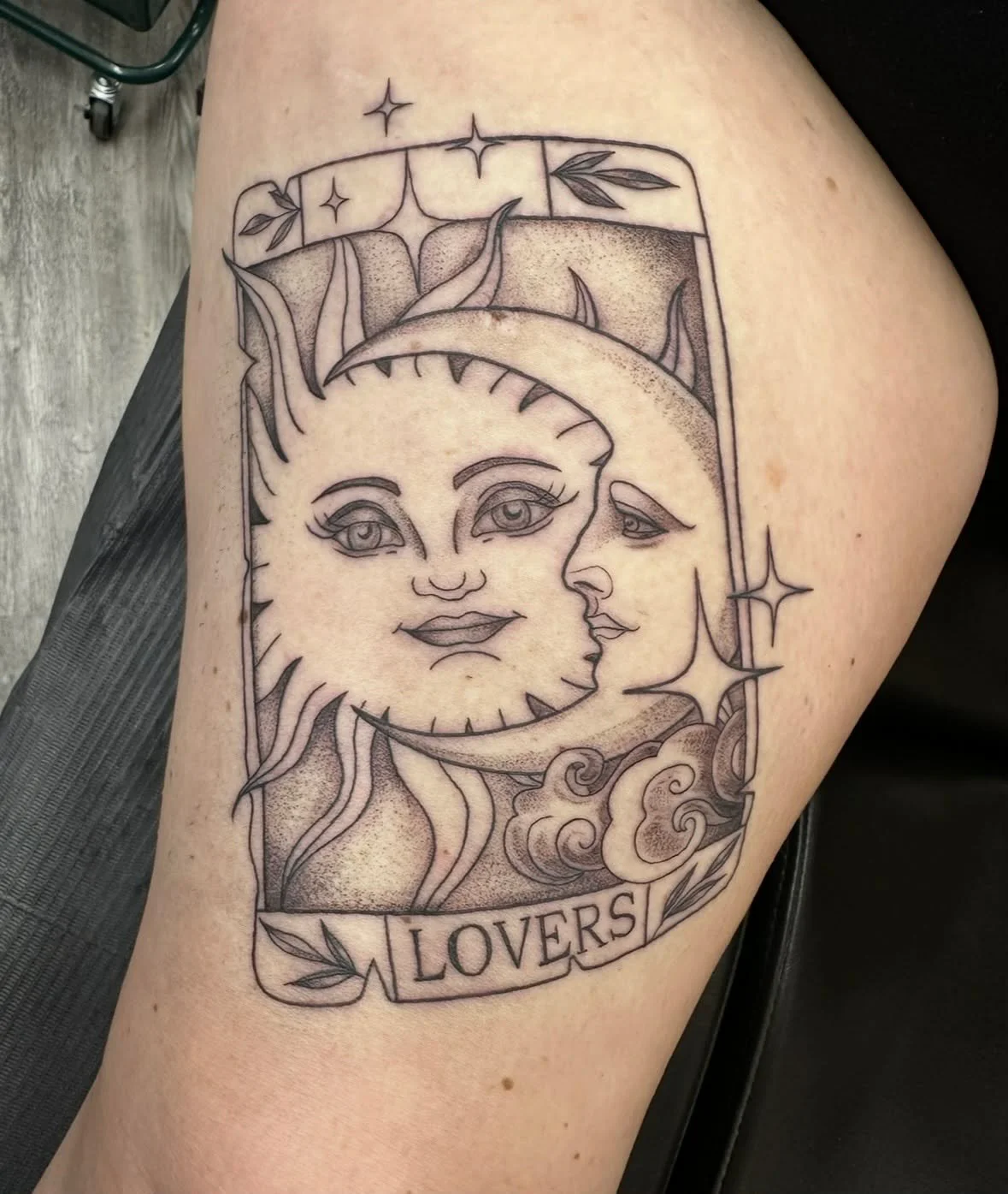 Tattoo of an illustrated sun and moon with faces, surrounded by clouds, stars, and leaves, with a banner reading "LOVERS."