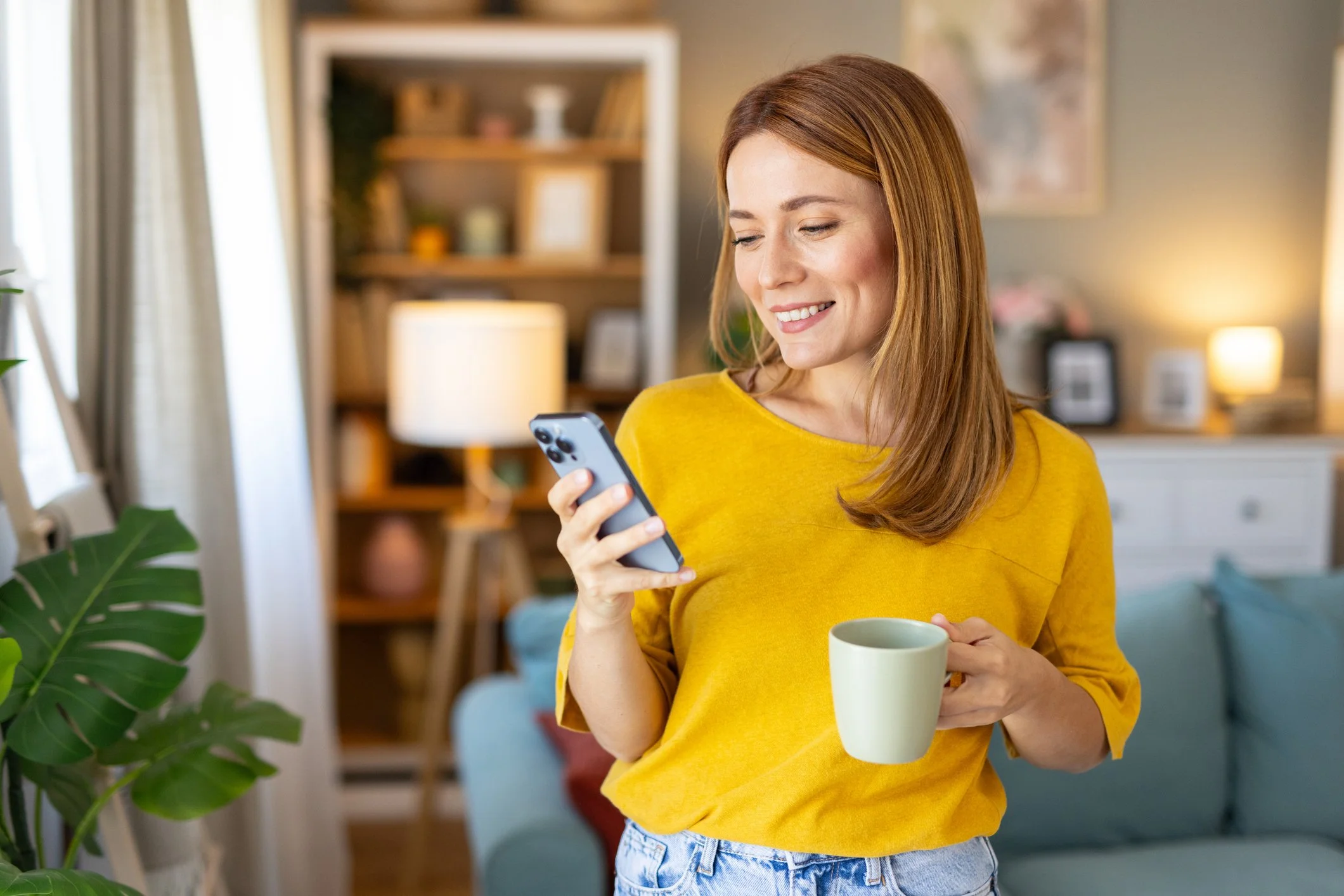 A woman with red hair in a yellow shirt holding a smartphone in one hand and a white mug in the other, standing in a cozy living room with a bookshelf, a lamp, and a sofa. Menopause, OGBYN, Obstetrics, Weight Loss