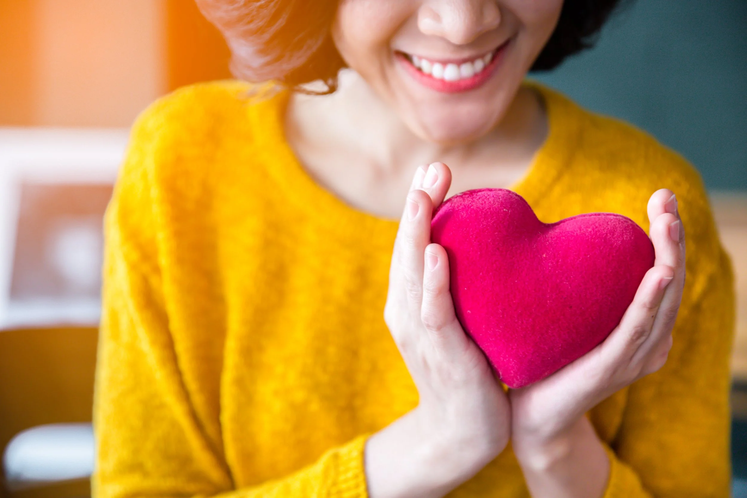 A woman in a yellow sweater holding a pink plush heart, smiling. Menopause, OGBYN, Obstetrics, Weight Loss.