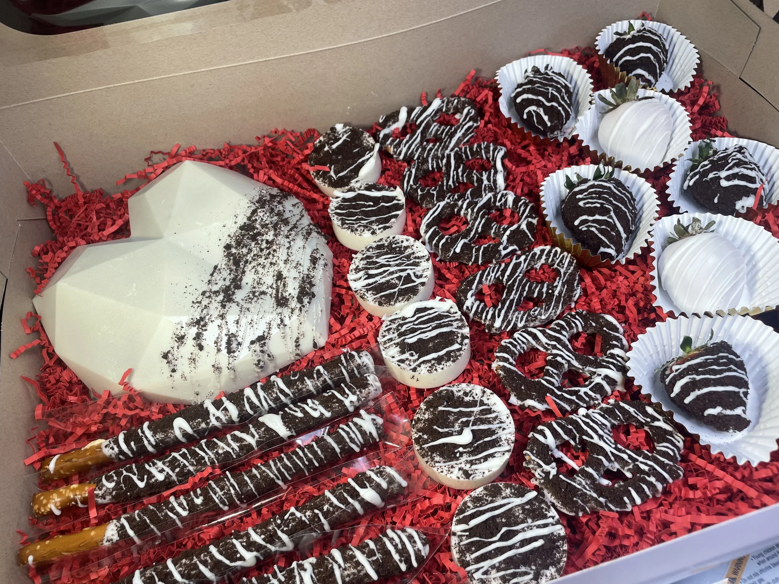 Chocolate and white-striped truffles, chocolate-covered pretzel rods, white chocolate dipped strawberries, and a white geometric heart-shaped cake in a box lined with red paper shred.