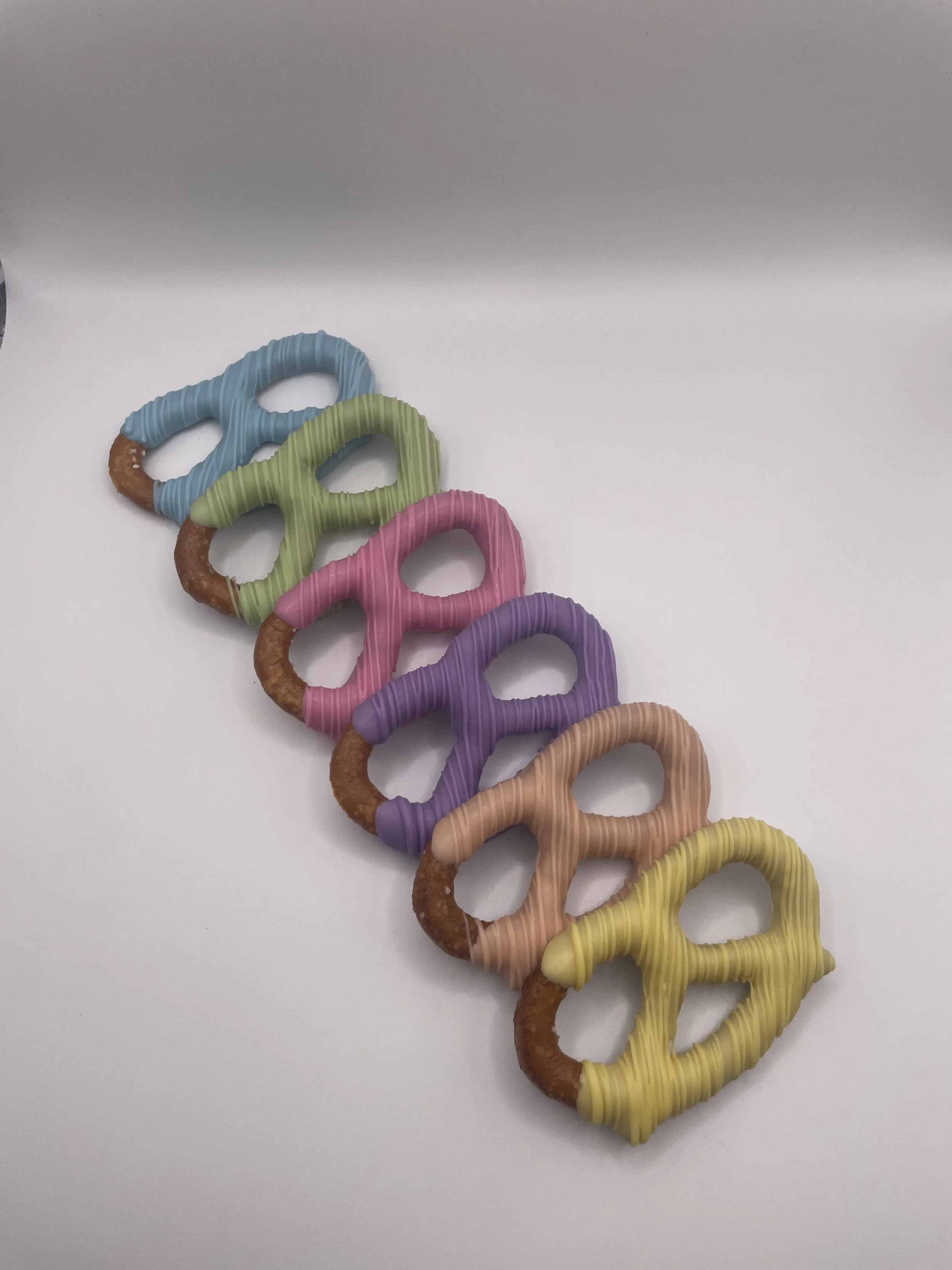 Colorful pretzels with pastel icing in blue, green, pink, purple, peach, and yellow, arranged in a diagonal line on a white surface.
