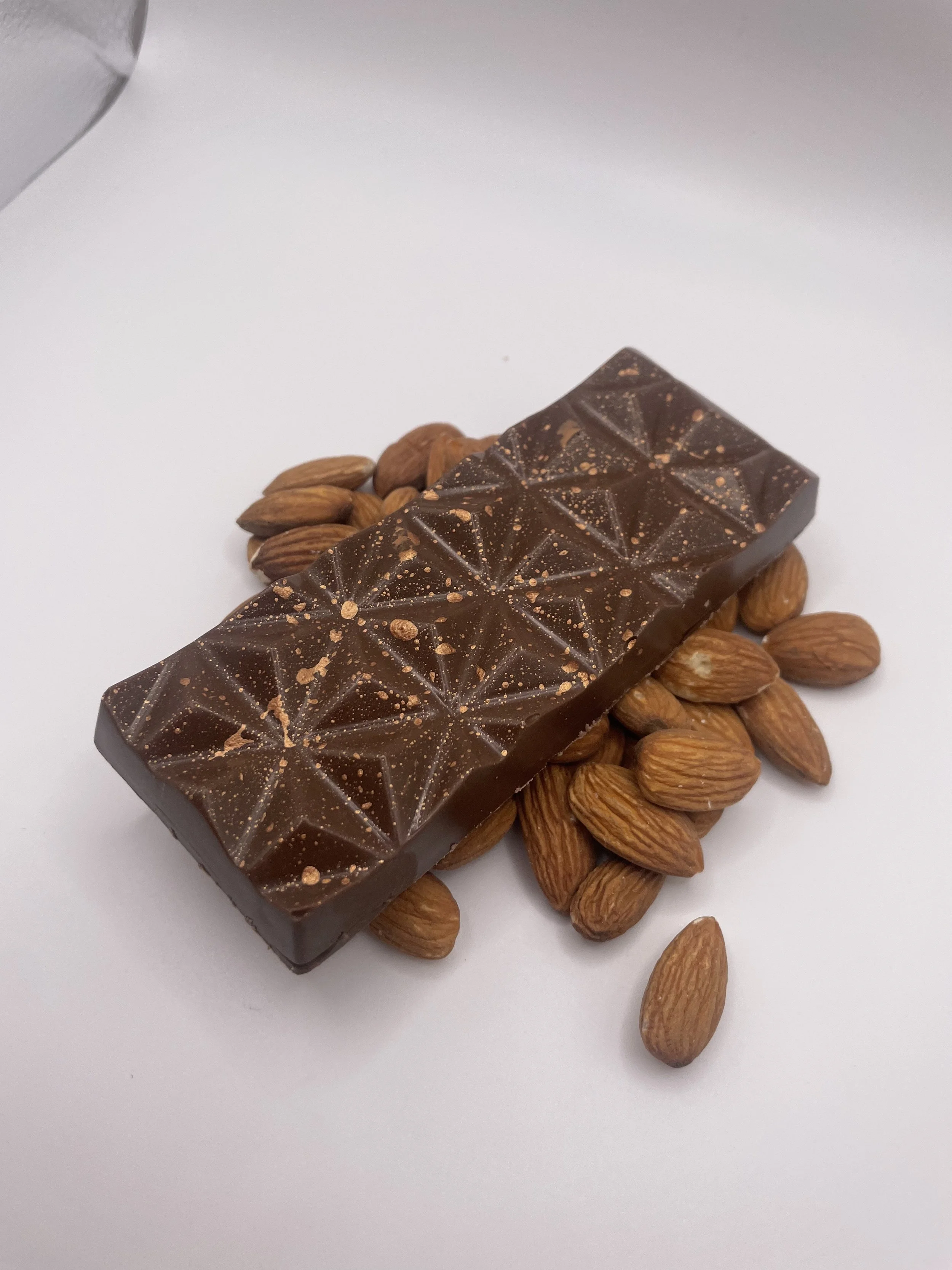 A chocolate bar with geometric patterns and small sprinkled particles, resting on a pile of almonds on a white surface.