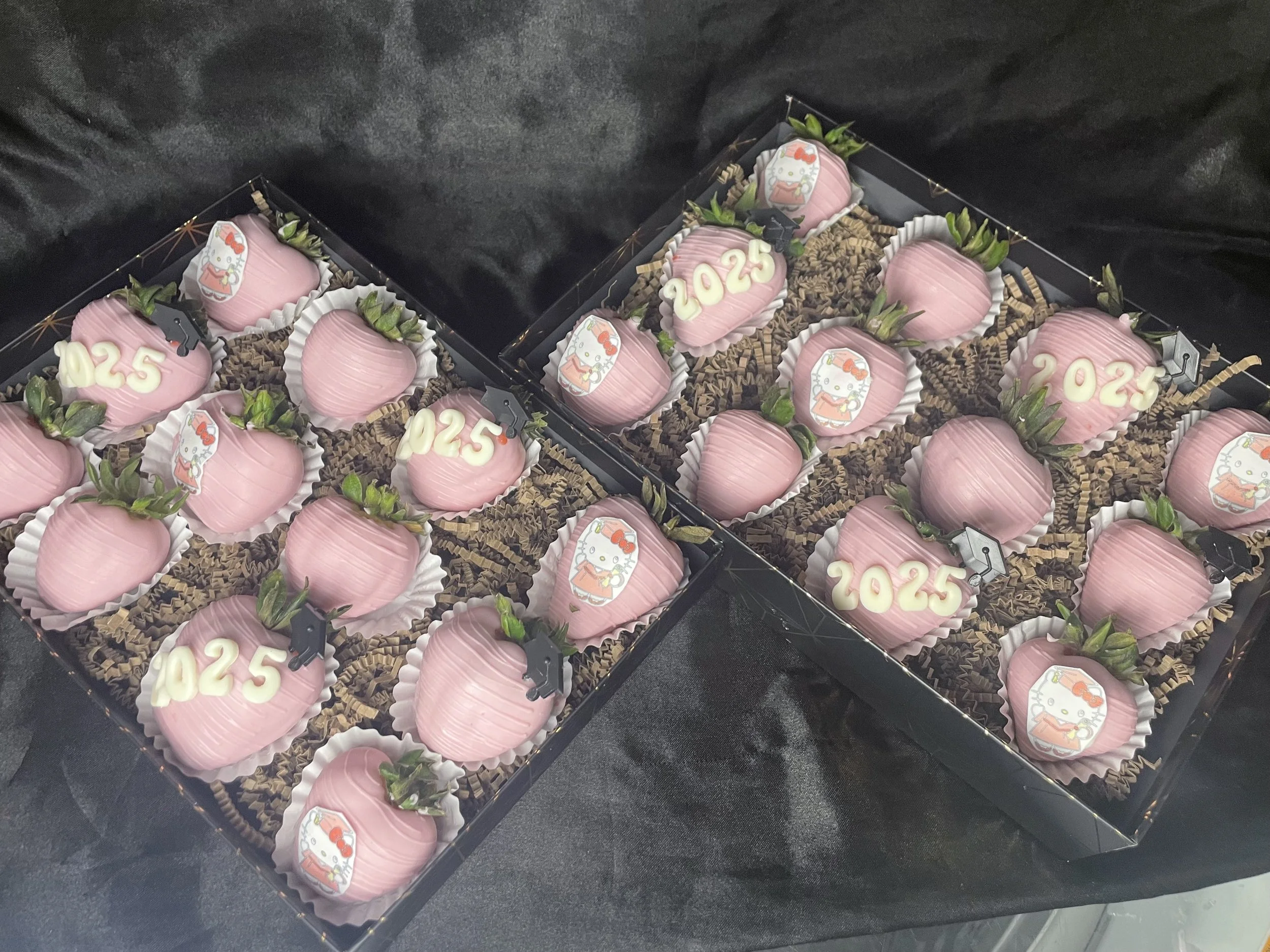 Two boxes of pink strawberries decorated with Hello Kitty images, the number 2025, and small black graduation caps, set on a black cloth background.
