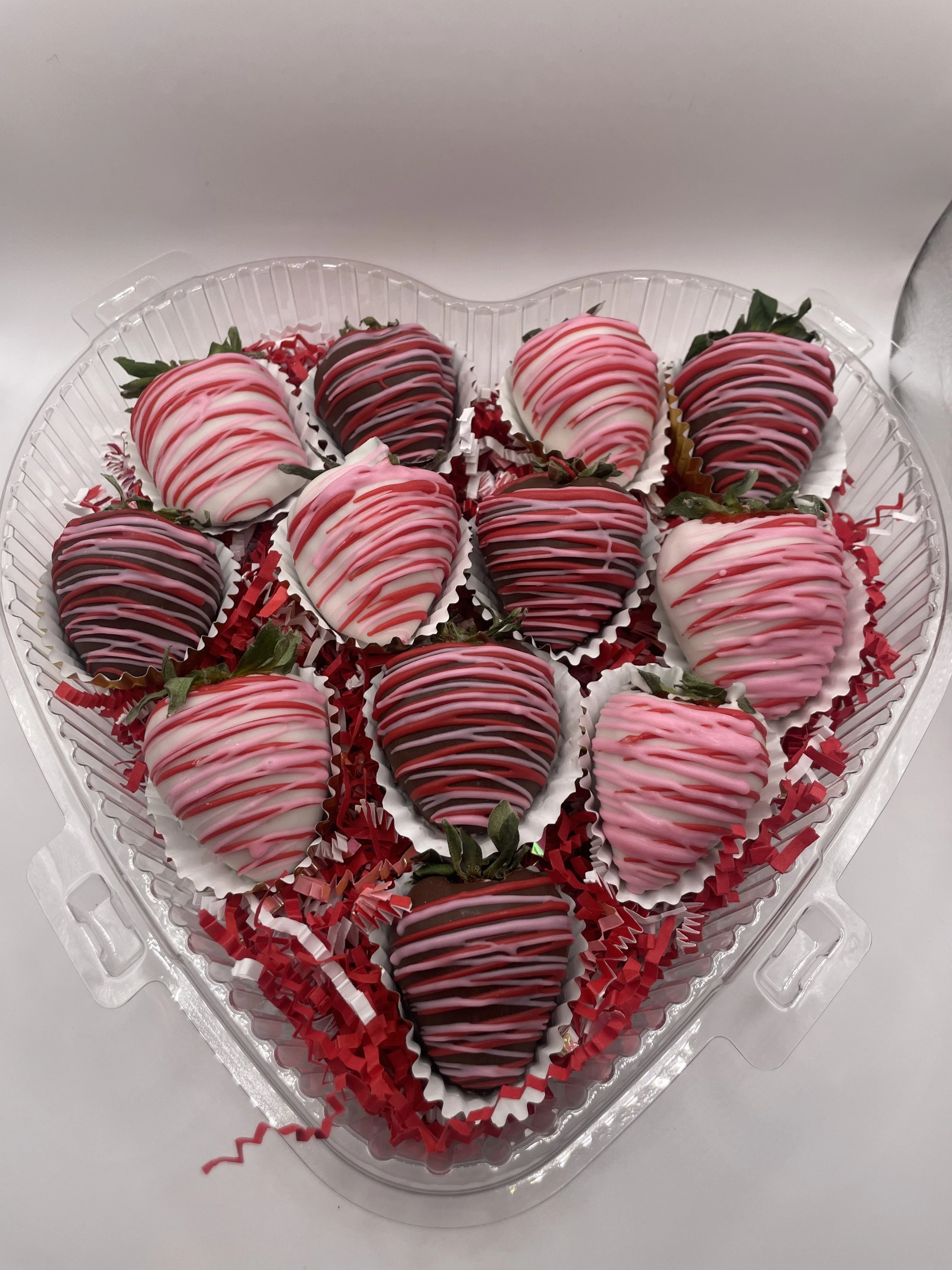 Heart-shaped plastic tray filled with decorated strawberries, some dark chocolate and some white chocolate, drizzled with pink and white icing.