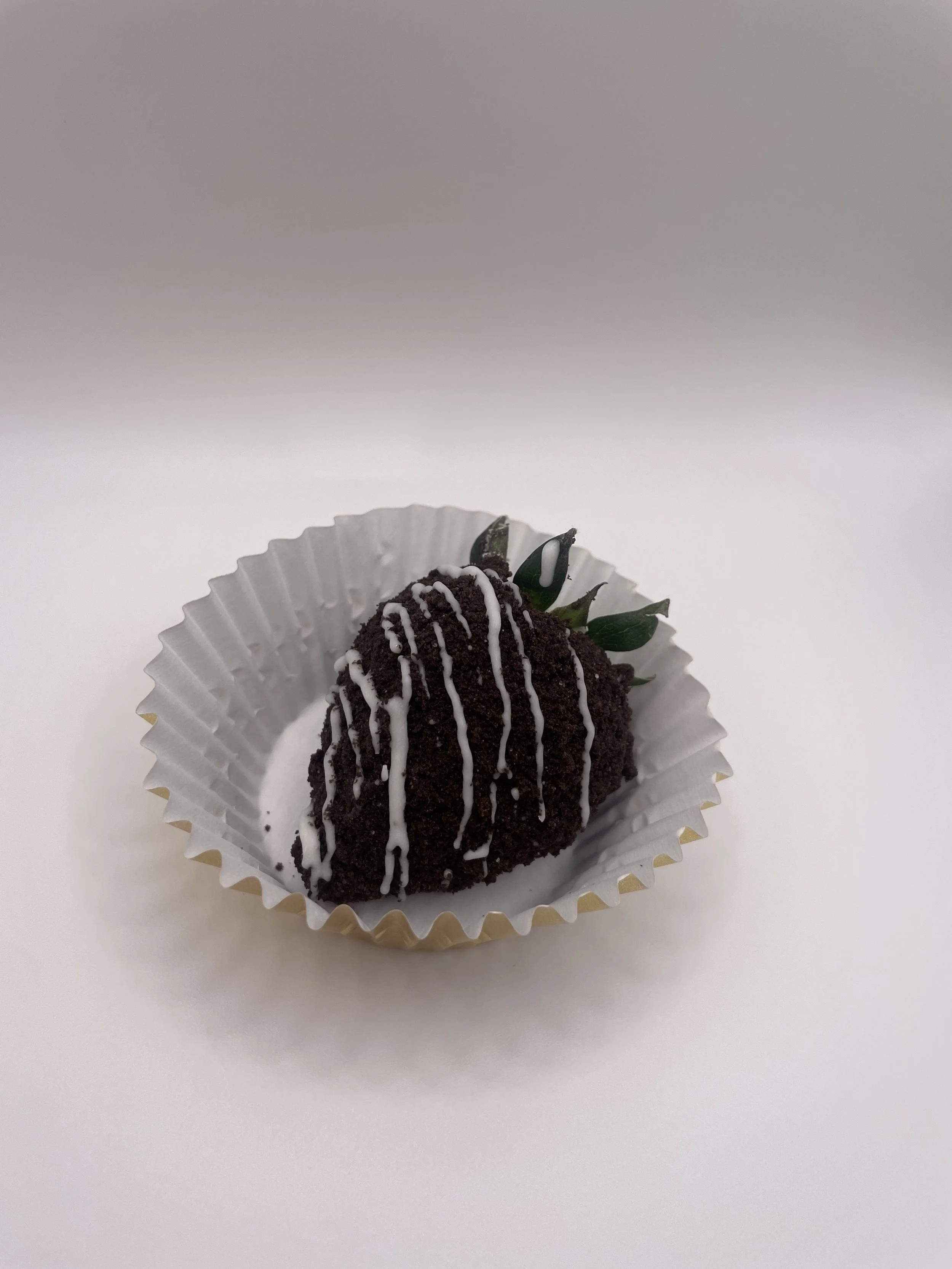 Chocolate cake with white icing drizzle, garnished with a green leaf, placed in a white paper cup on a white surface.