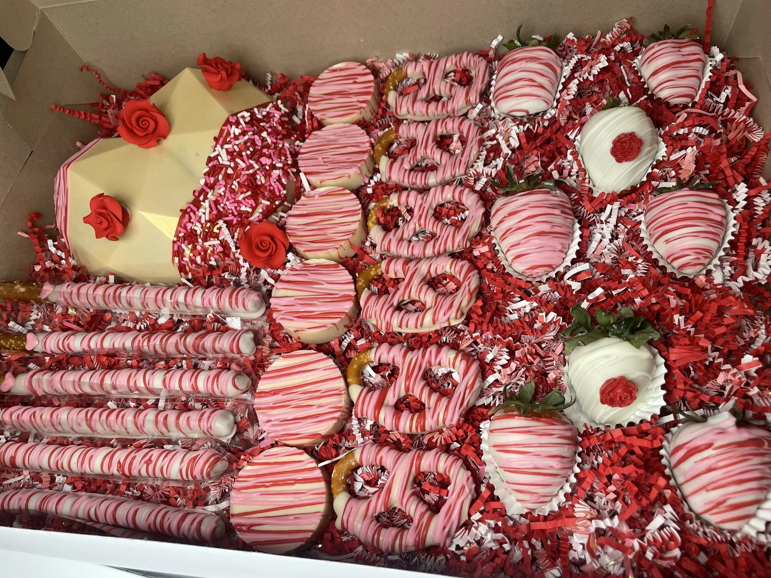 Assorted Valentine's Day chocolates and treats decorated with red, white, and pink stripes, including heart-shaped, round, and elongated confections, placed on shredded red and white paper in a box.