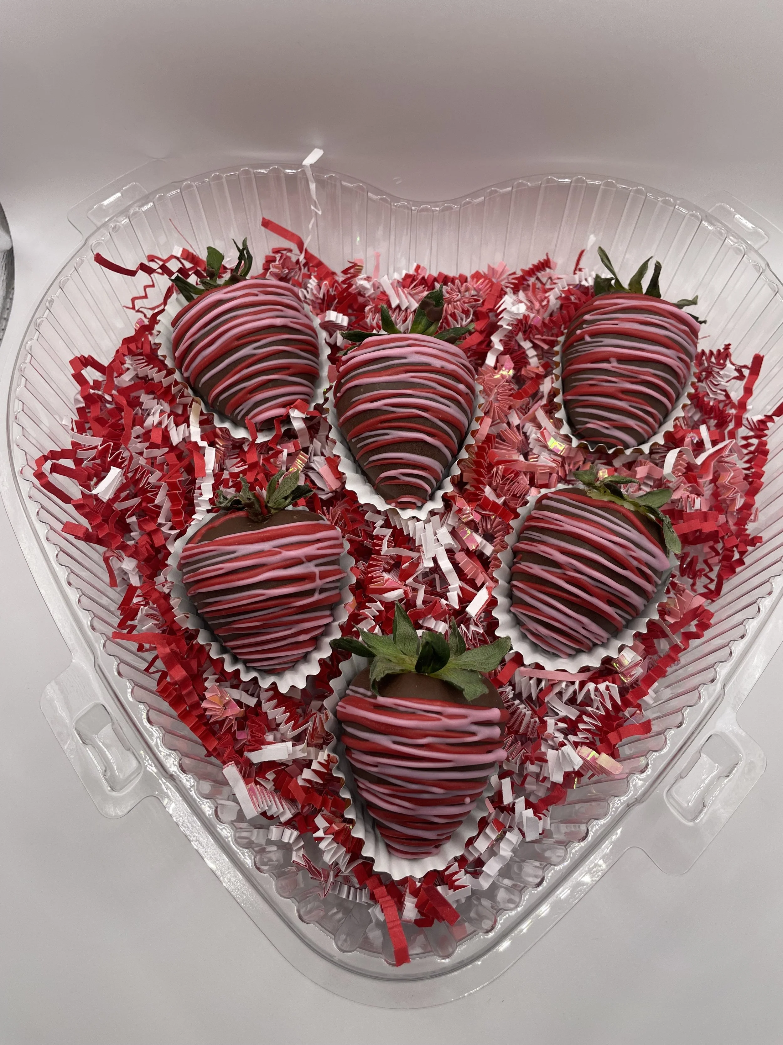 Chocolate-covered strawberries decorated with pink and white icing drizzle inside a clear plastic container lined with red and white shredded paper.