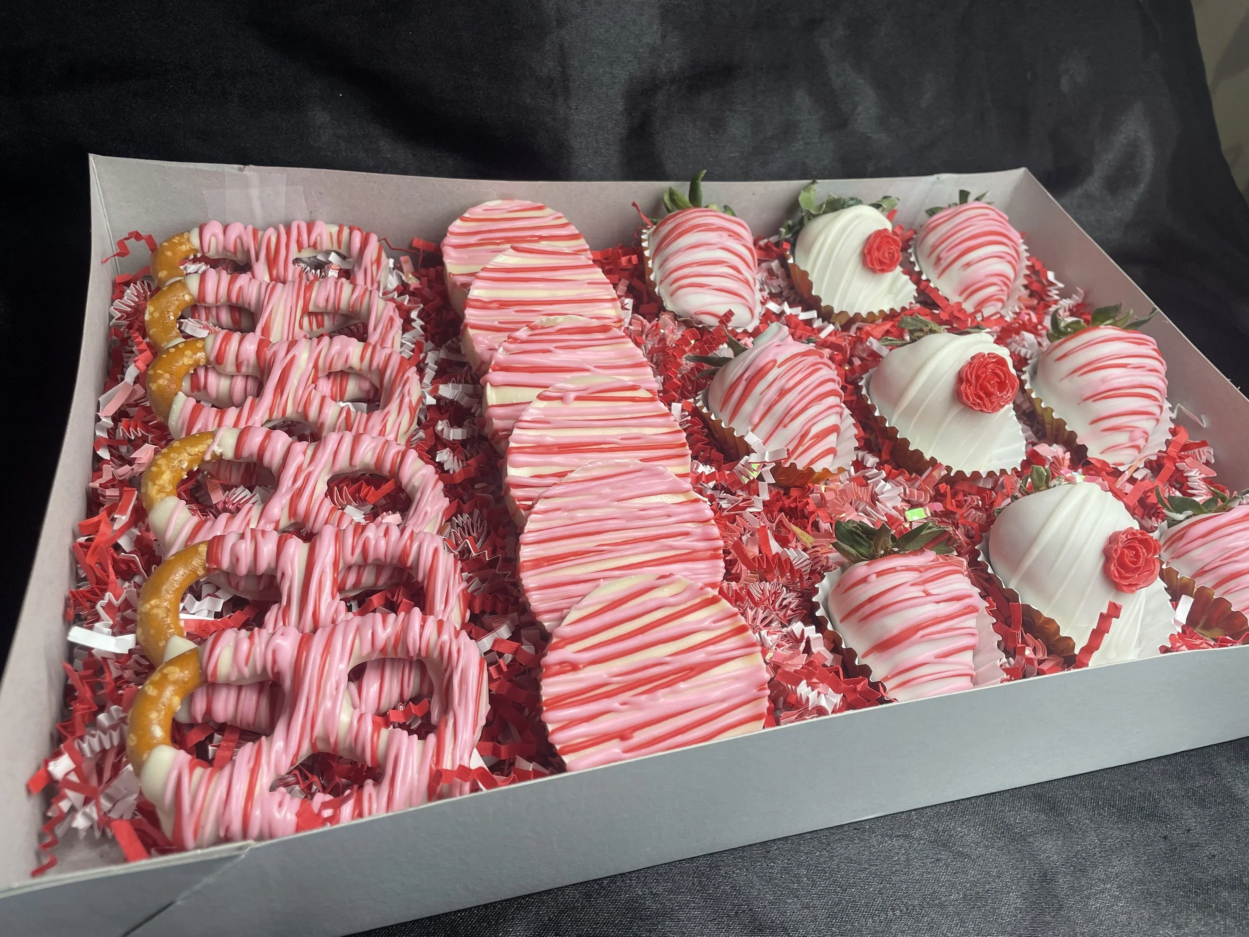 Box of decorated strawberries and pretzels with pink and white drizzle, arranged on red shredded paper.