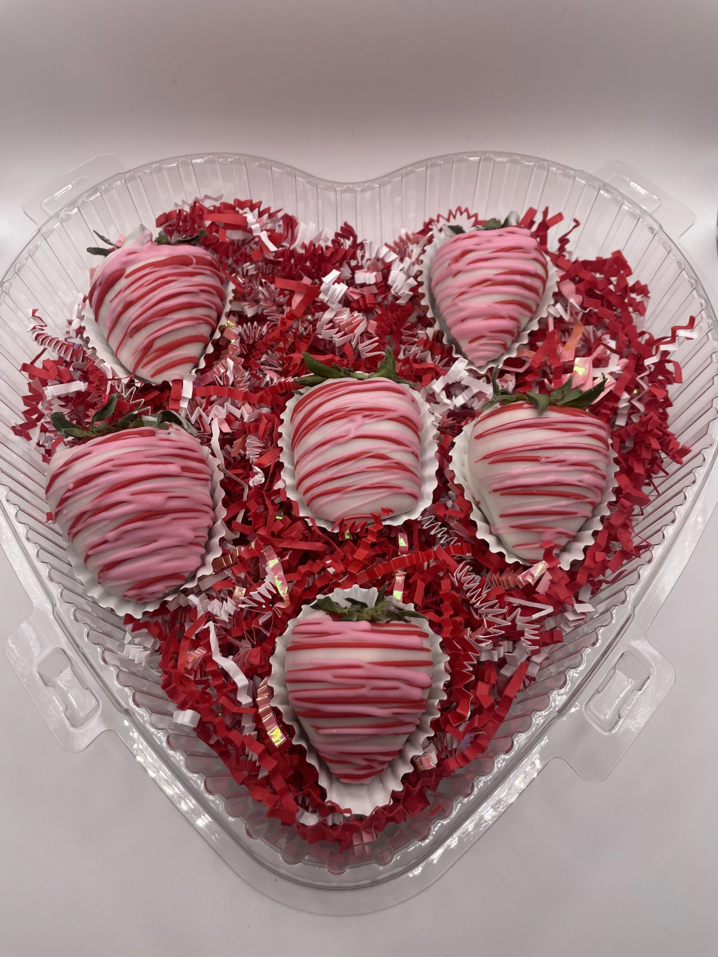 Heart-shaped container with six white chocolate strawberries drizzled with pink and red chocolate, surrounded by red and white decorative shredded paper.