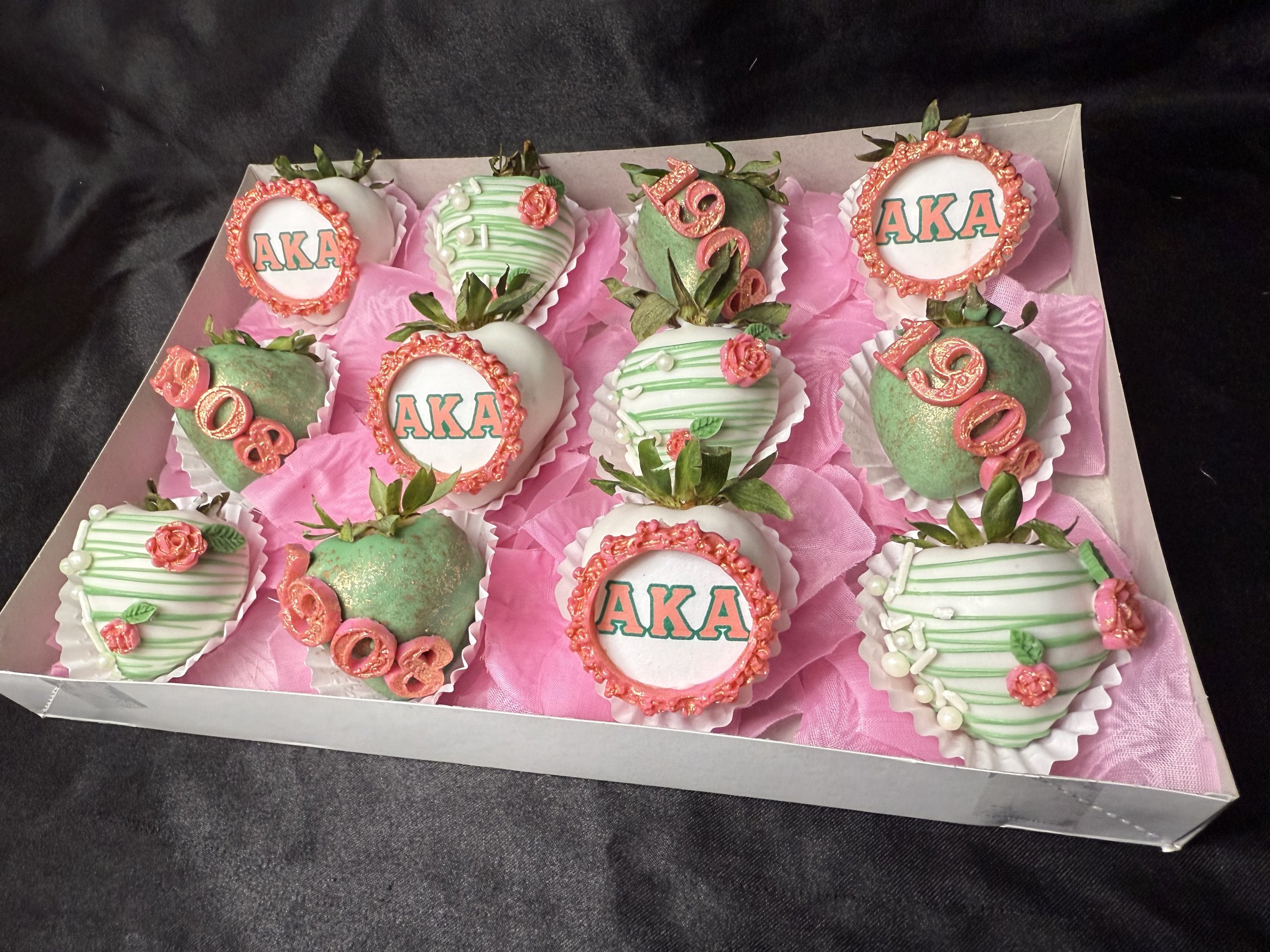 Box of decorative strawberries for sorority celebration, some with 'AKA' and '1908' written on them, decorated with pink, green, and white icing.