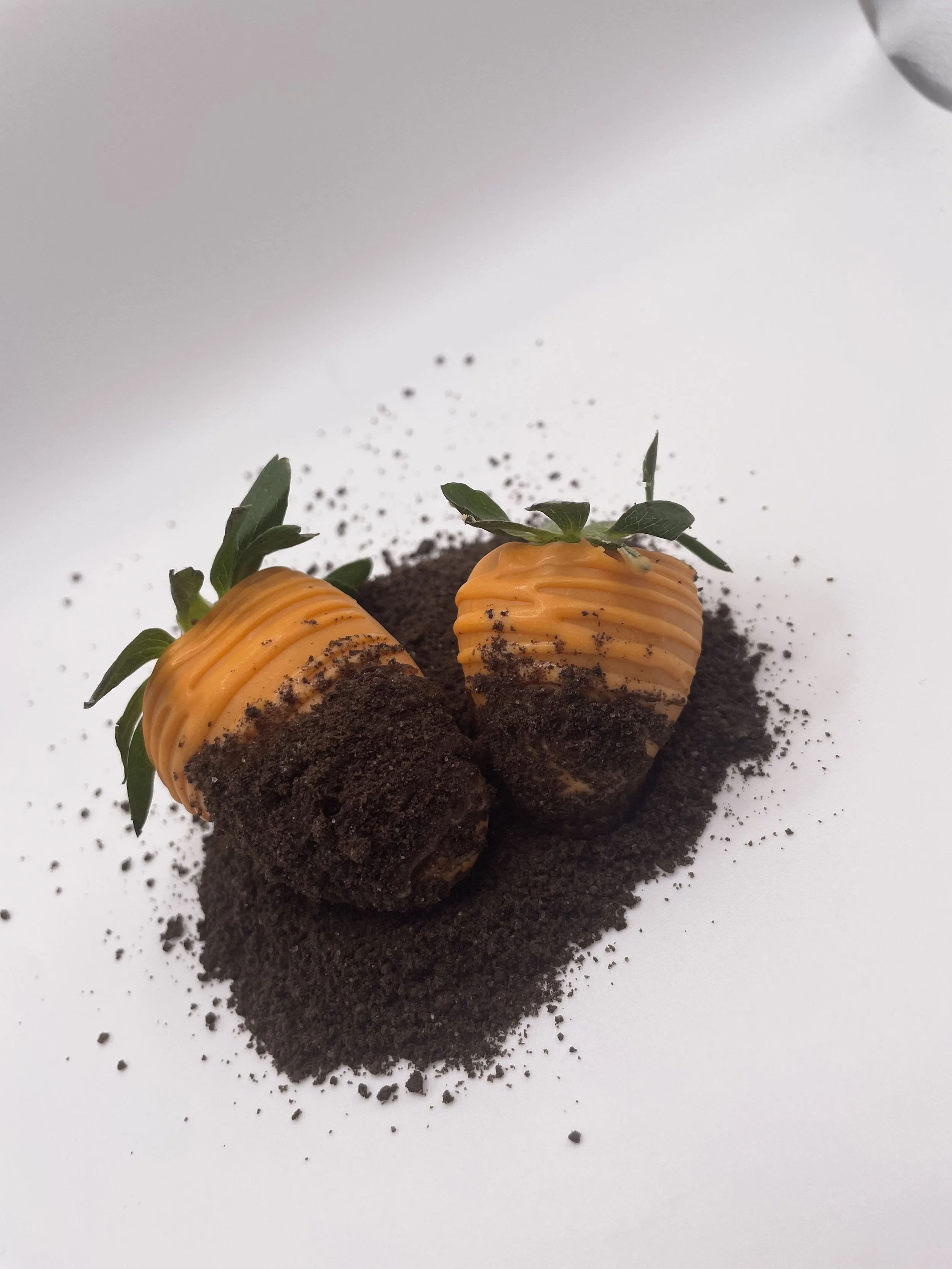 Two strawberries wrapped in orange-colored chocolate, garnished with green leaves, placed on a bed of crushed black cookie crumbs.