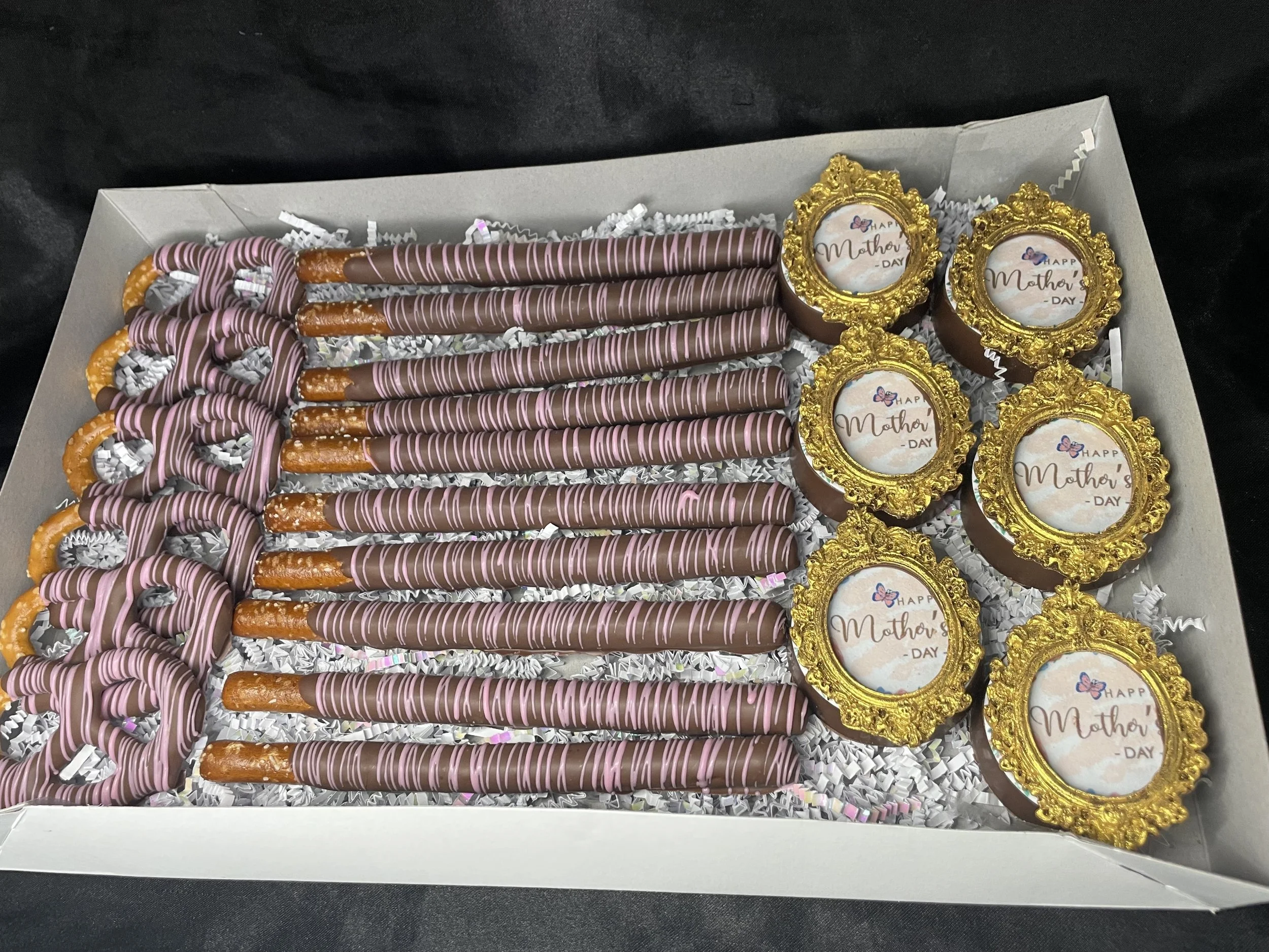 Box of Mother's Day themed chocolates, including gold framed chocolates with 'Happy Mother's Day' printed on them, pink and brown striped pretzel sticks, and pink and brown striped chocolate-covered pretzels, arranged with white crinkle paper.