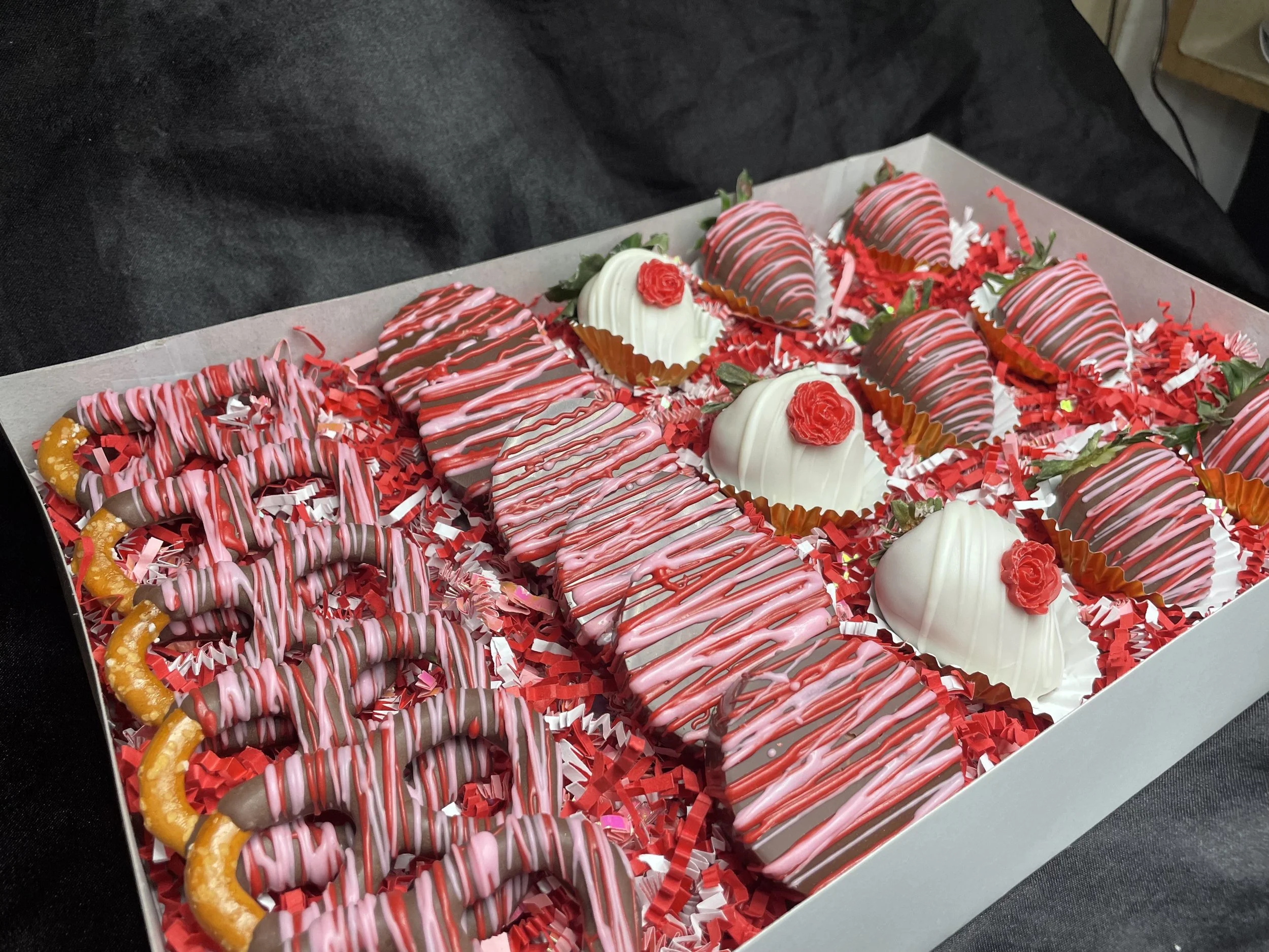 A white box filled with decorated cookies and desserts on red shredded paper, including strawberry-shaped cookies, white domed desserts with red roses, and variously decorated cookies with pink and white icing.