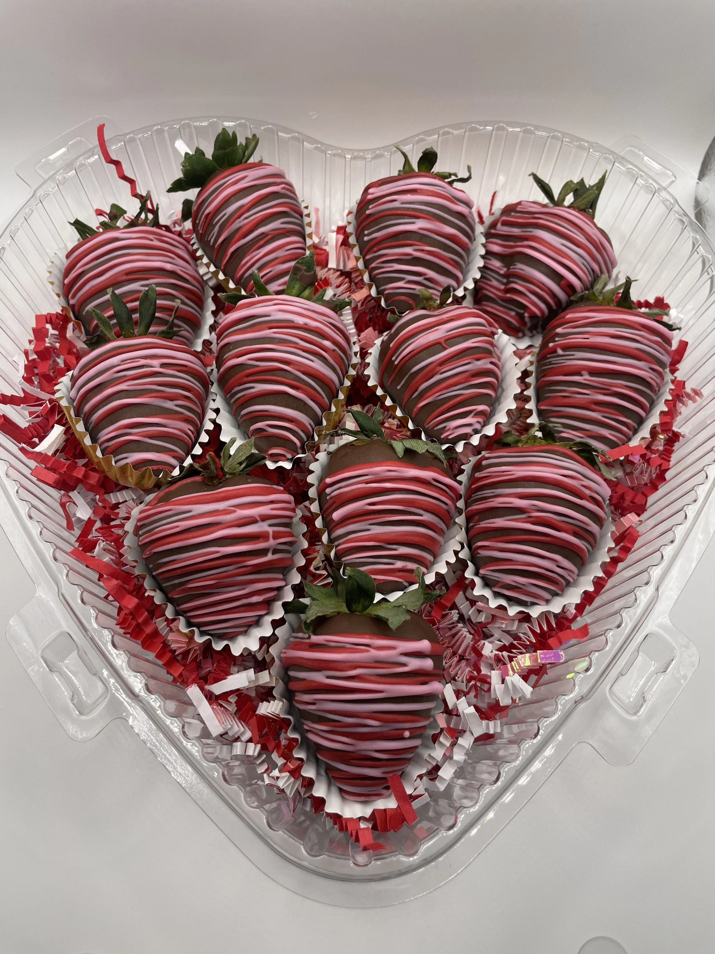 A heart-shaped plastic container filled with decorated strawberries with pink and white drizzle, placed on a white surface.