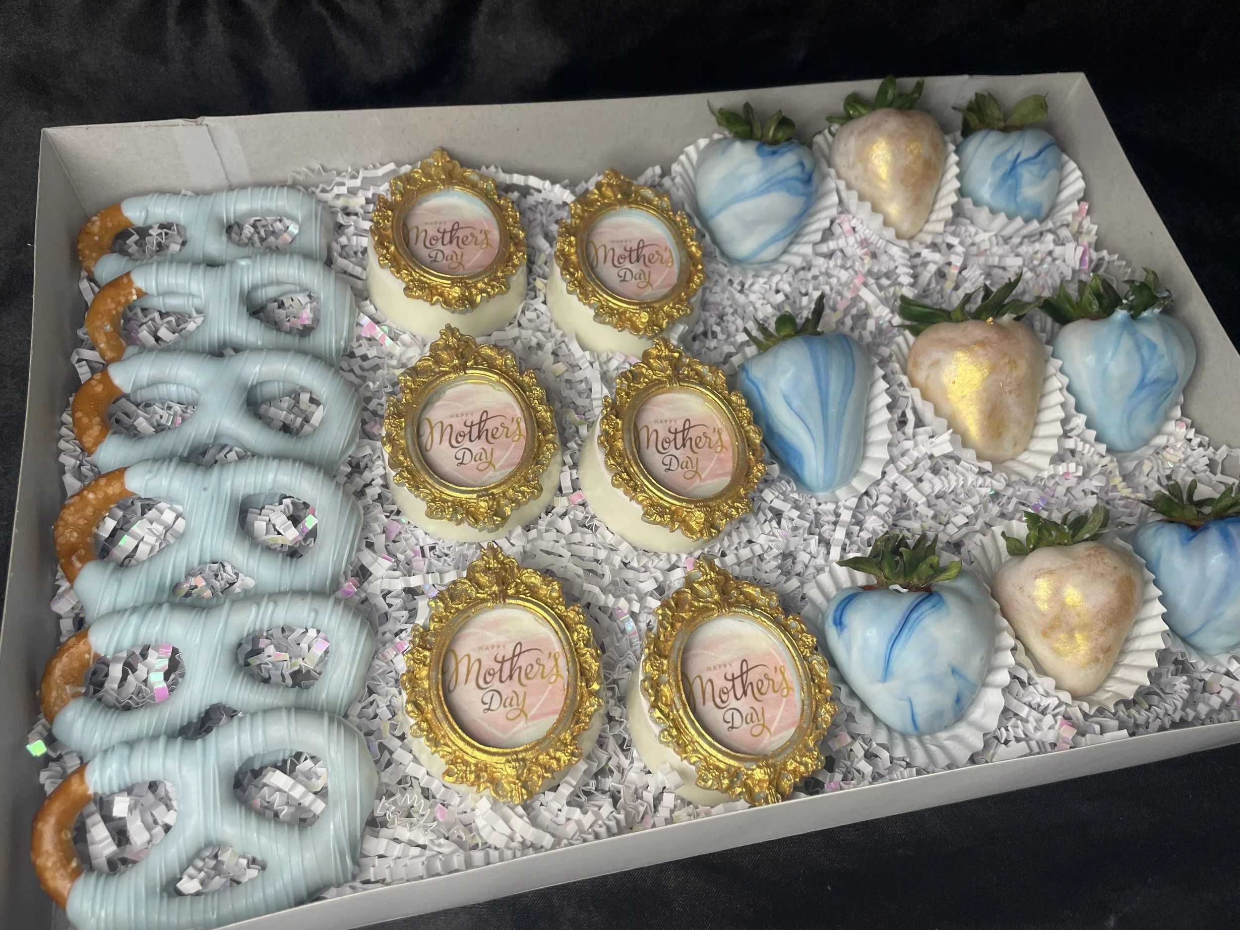 A box of decorated cookies and chocolates for Mother's Day, featuring pretzels with blue icing, chocolate-covered strawberries, and edible gold accents.