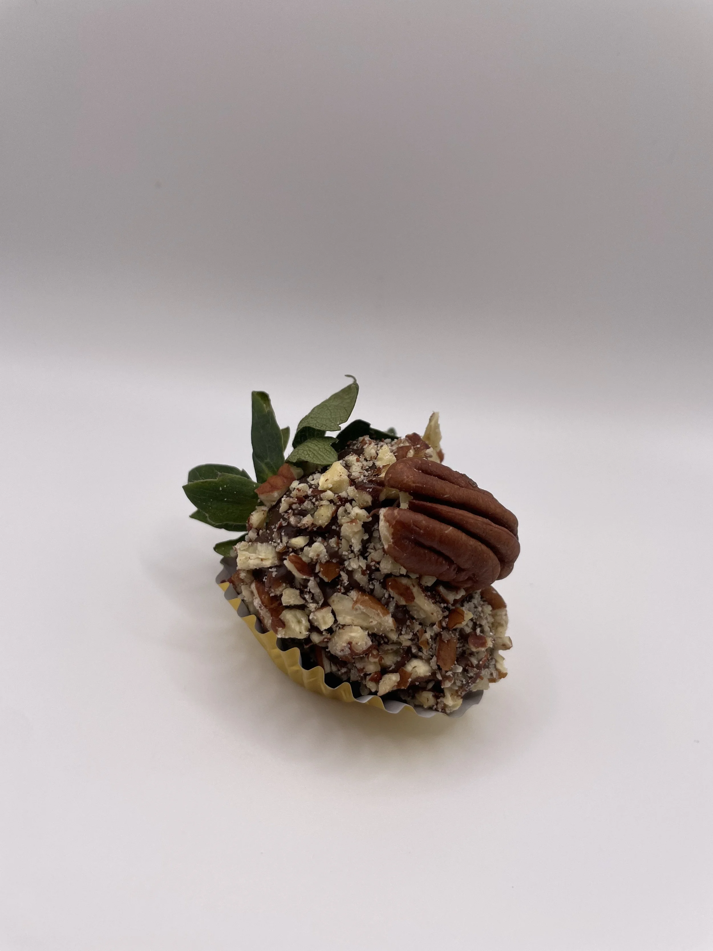 Chocolate truffle coated with chopped nuts, topped with a pecan half and green leaves, on a white background.