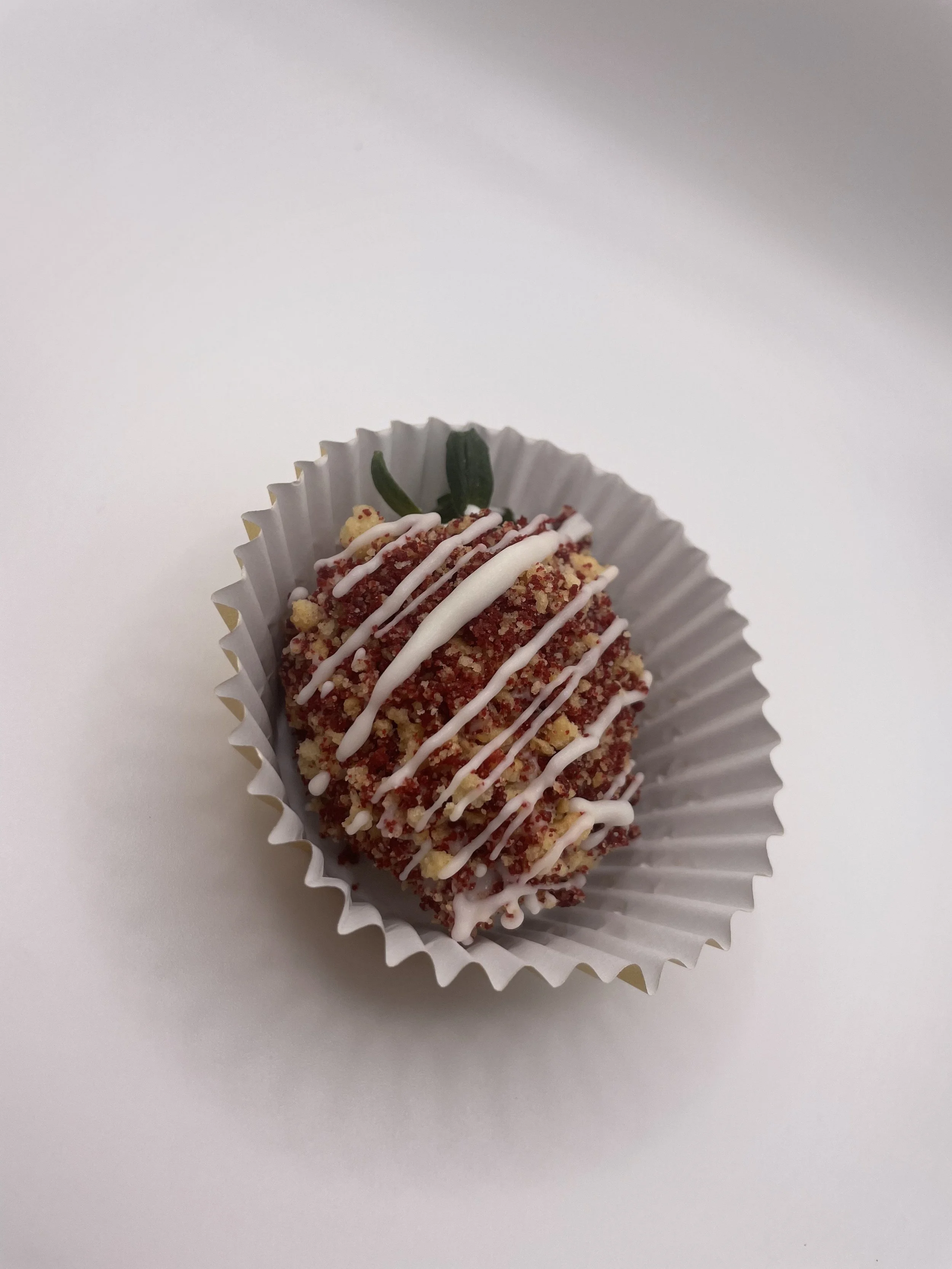 A chocolate confectionery covered with red crumbs, white icing drizzle, and a small green decorative topping, placed in a white paper cup.