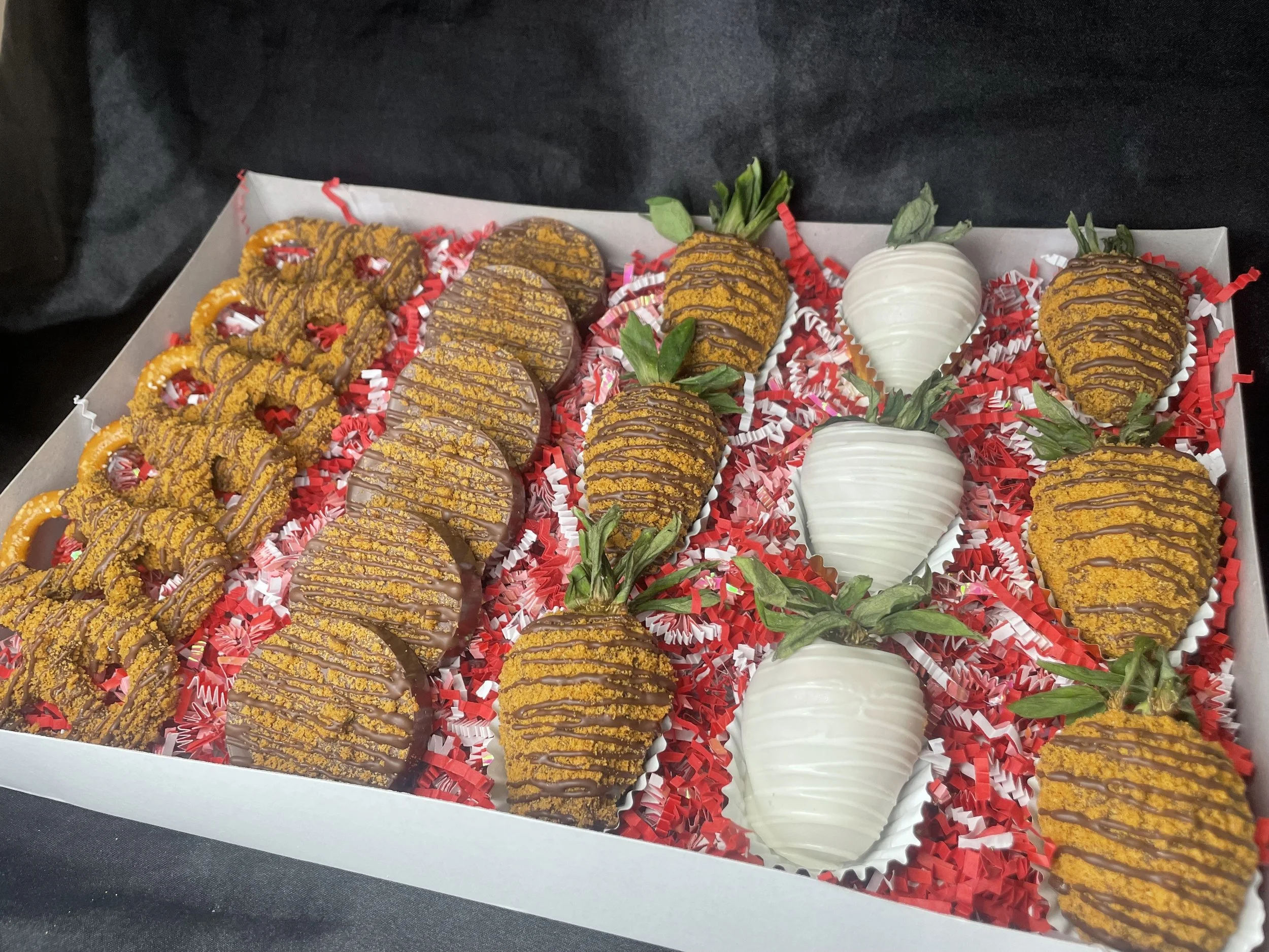 A box of decorated strawberries, some dipped in white chocolate and others with chocolate and golden sprinkles, arranged on red and white shredded paper.
