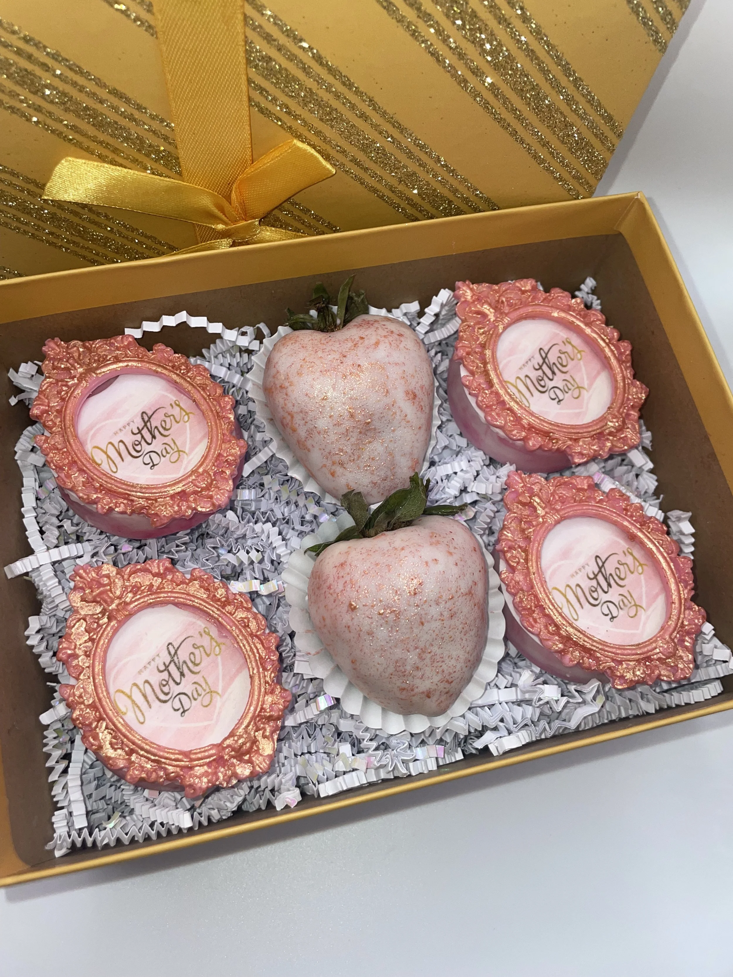 A gift box containing two decorated strawberries and four heart-shaped chocolate candies with 'Mother's Day' written on them, surrounded by white shredded paper.