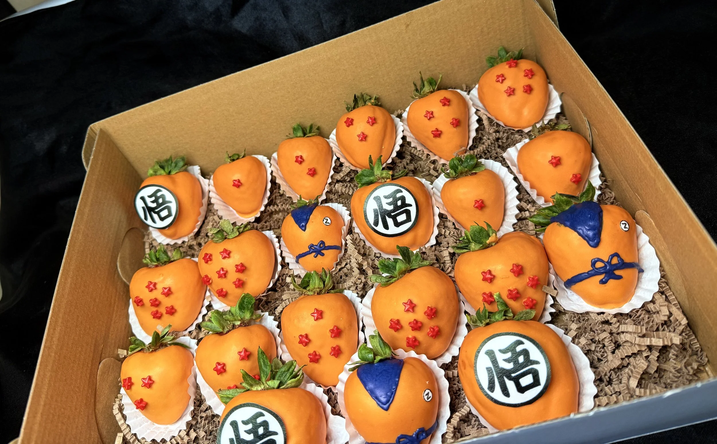 A box of artificial strawberries decorated with orange icing, red star-shaped sprinkles, and some with blue and white decorations resembling Dragon Ball characters.