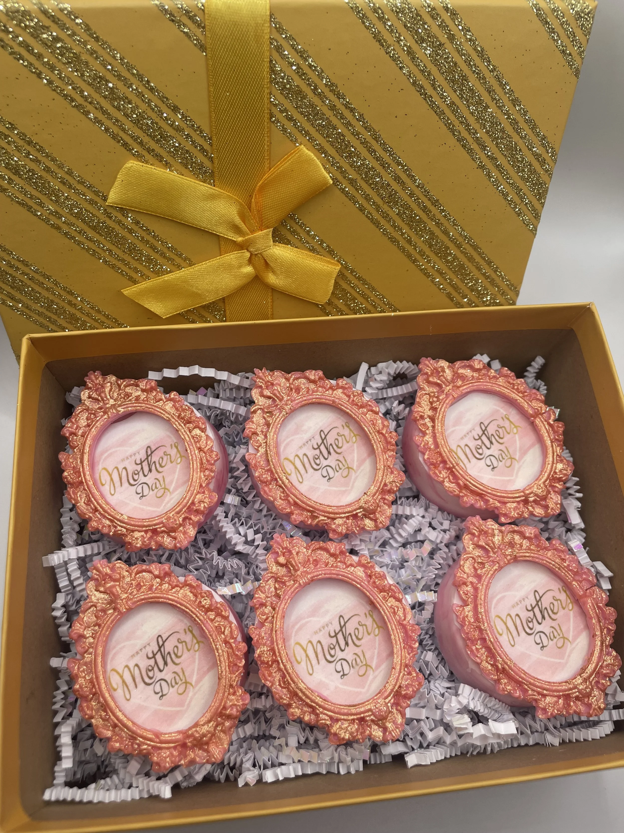 Six decorative cookies in a box, each with a pink and gold ornate oval frame and the words 'Happy Mother's Day' written in gold. The box has a gold glittery pattern and a yellow ribbon tied in a bow at the top.