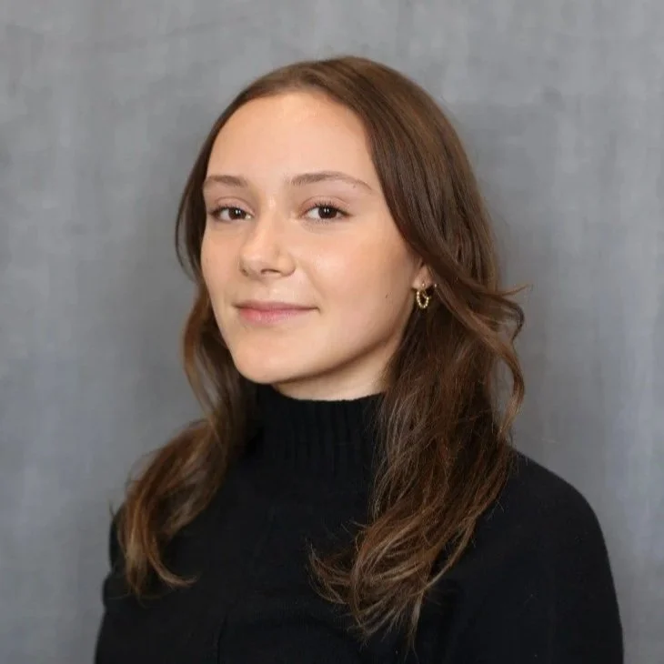 A young woman with light skin and brown, wavy hair, wearing small hoop earrings and a black turtleneck sweater, standing against a gray background.