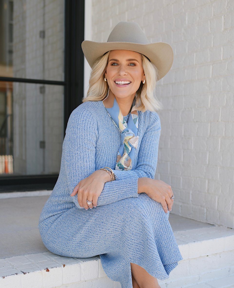 A woman with blonde hair wearing a wide-brimmed hat, blue sweater, patterned scarf, and earrings, sitting on a white ledge outside near a brick wall, smiling at the camera.