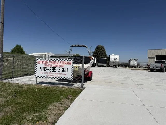 Boat on a trailer parked at Hot Rods Vehicle Storage, a climate-controlled storage facility with RVs and trucks visible in the background on a sunny day.