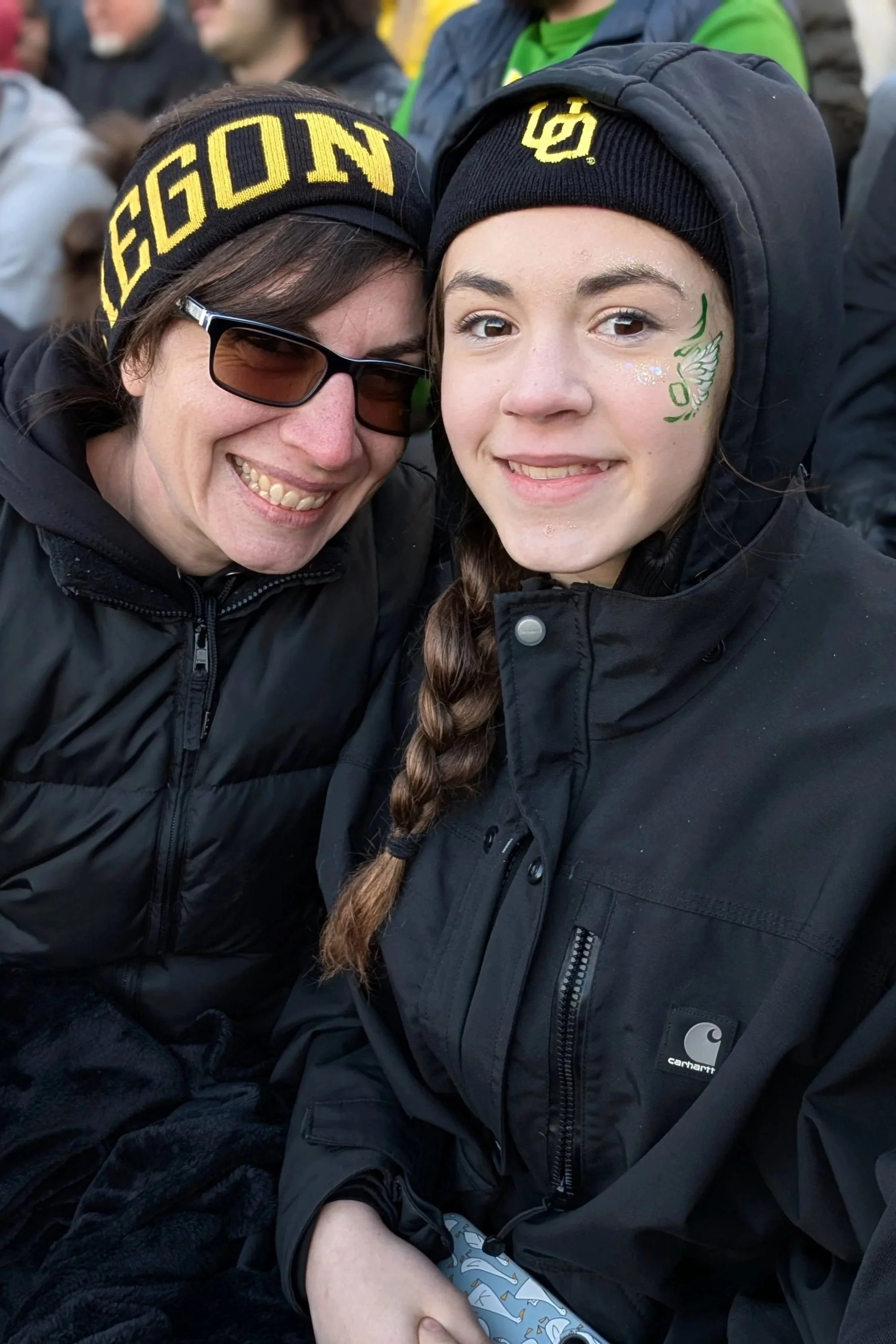 Two women dressed in black jackets and hats, smiling closely together. One woman wears sunglasses and a beanie with the word 'BEGON' on it, while the other has face paint and a Carhartt jacket, in a crowded outdoor setting.