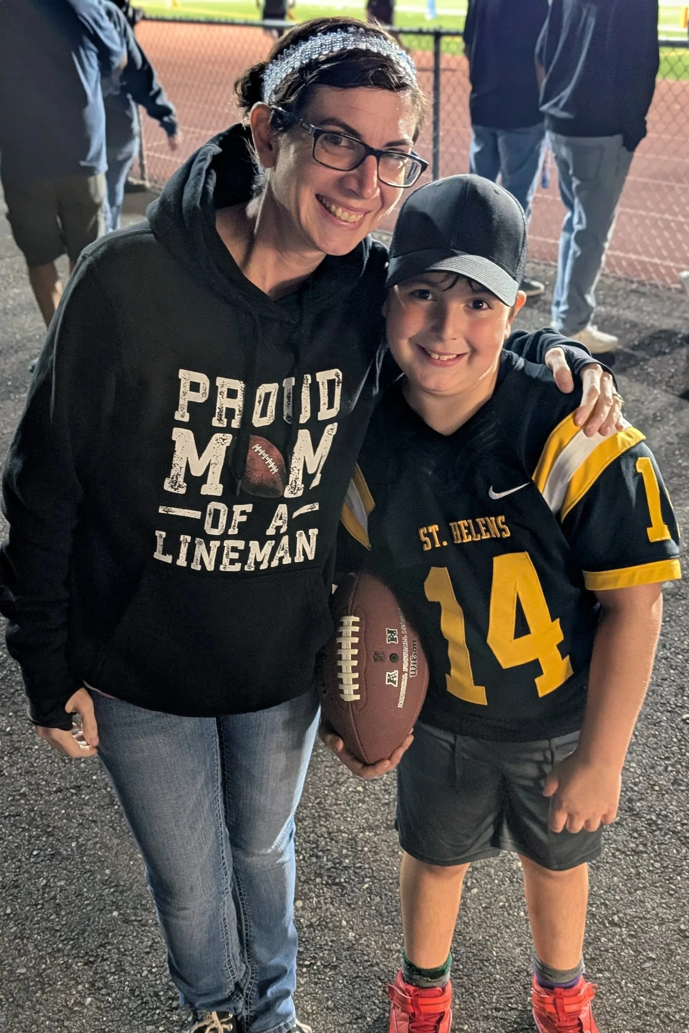 A woman and a young boy standing together on a sports field at night. The woman is wearing glasses, a black hoodie with the words "Proud Mom of a Lineman" and a football graphic, and jeans. The boy is in a black and yellow football uniform with the number 14, holding a football, and wearing a cap. They are smiling and hugging, with a chain-link fence and a track in the background.