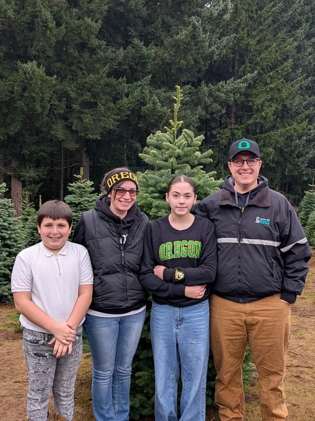 Family of four standing outdoors among Christmas trees in a forest, wearing casual clothing and displaying smiles.