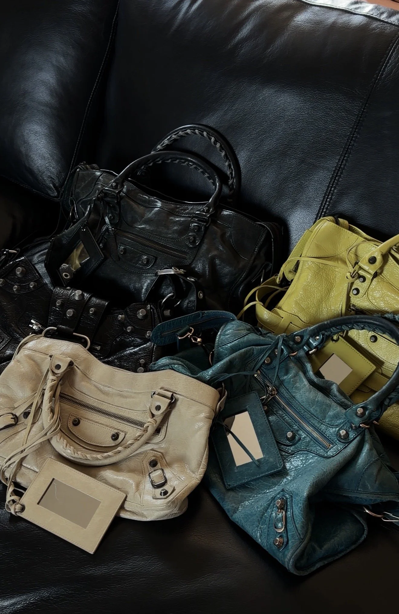 Multiple leather handbags in black, yellow, cream, and teal colors placed on a black leather couch.