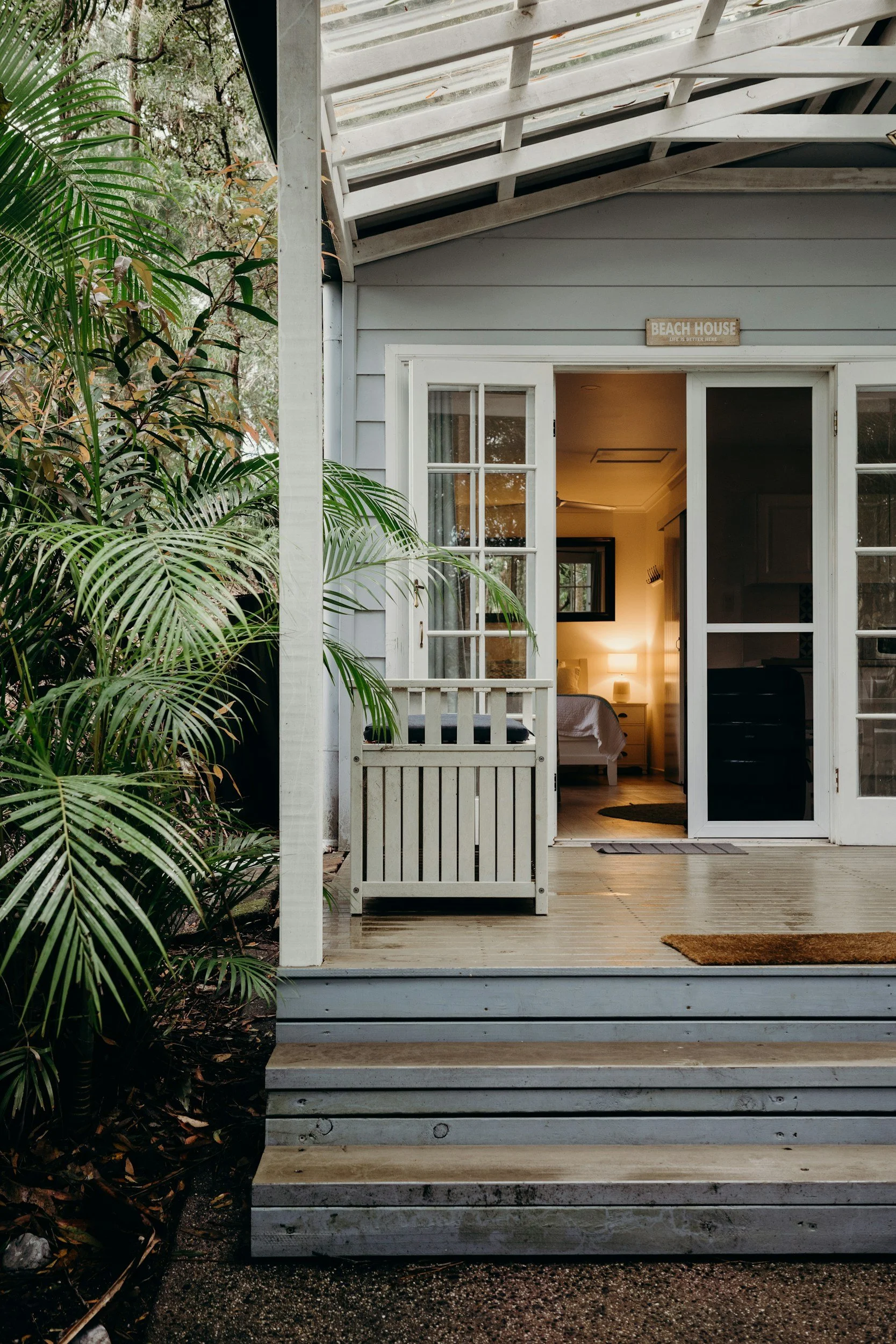 A wooden deck with steps leading to a door of a beach house, showing a bedroom with warm lighting inside. There are green plants on the left and a small sign above the door that reads 'Beach House'.