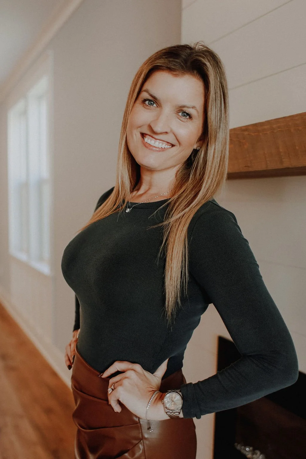 A woman with long, light brown hair and blue eyes, smiling while standing indoors. She is wearing a fitted black long-sleeve top, a watch, a bracelet, a ring, and a necklace, with her hand on her hip, in a room with light-colored walls and a wooden beam.