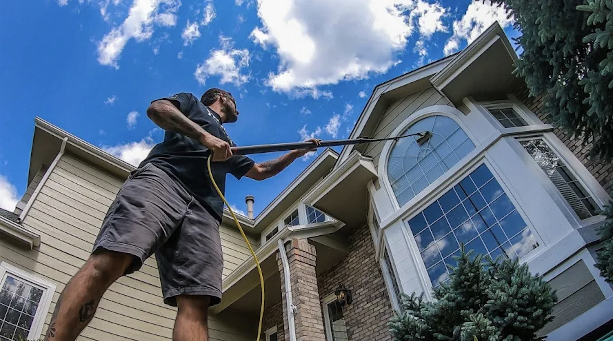A person using a pressure washer to clean the exterior of a house, which has large windows and a mix of siding and brick. The sky is blue with some clouds.