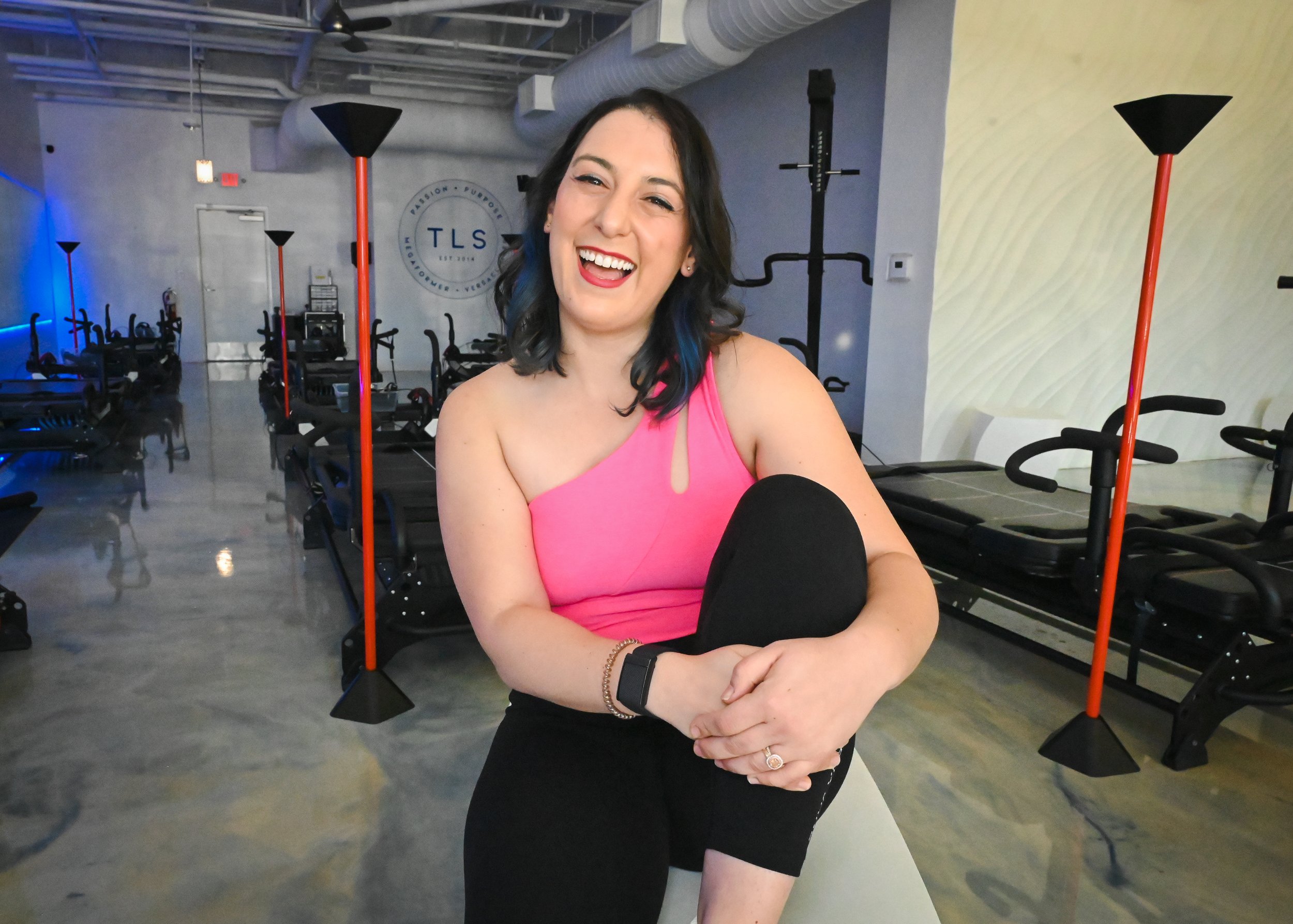 Smiling woman in a gym studio wearing a pink top, seated with leg crossed, surrounded by fitness equipment and mirrors.