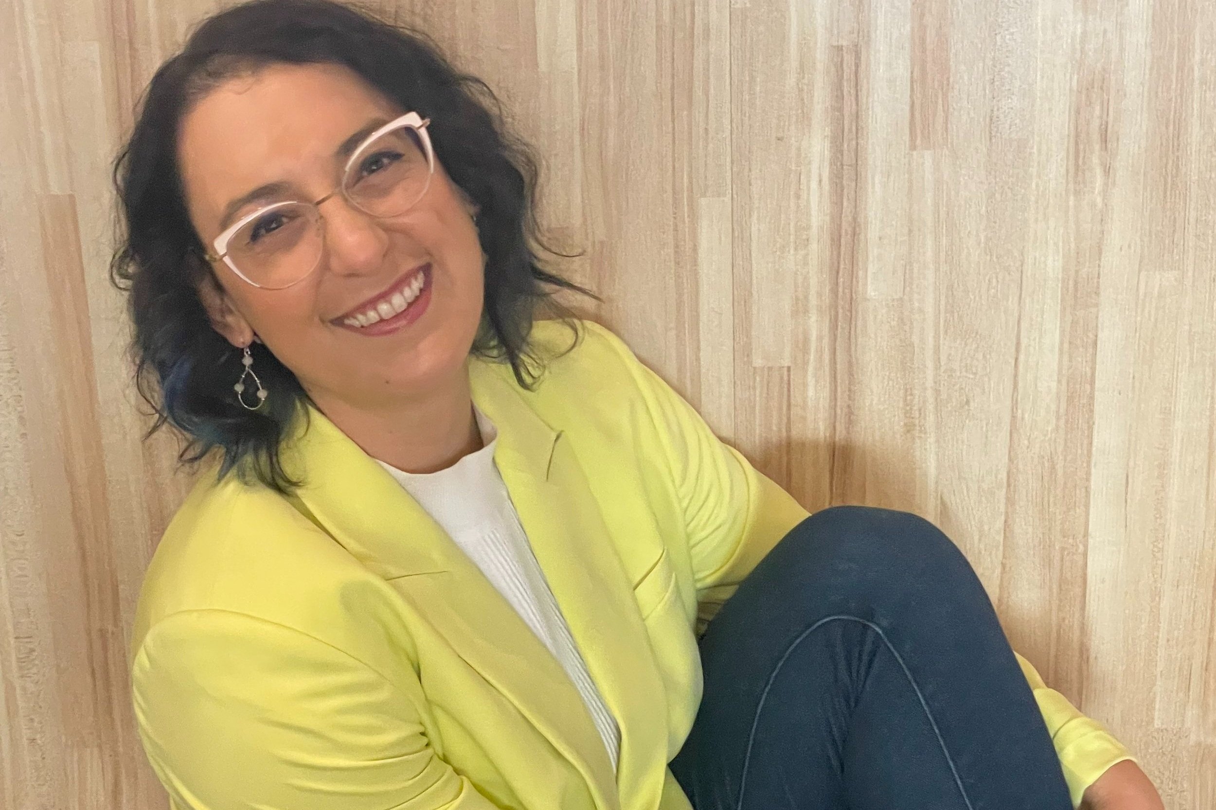 Smiling person wearing glasses and a yellow jacket sitting against a wooden background.
