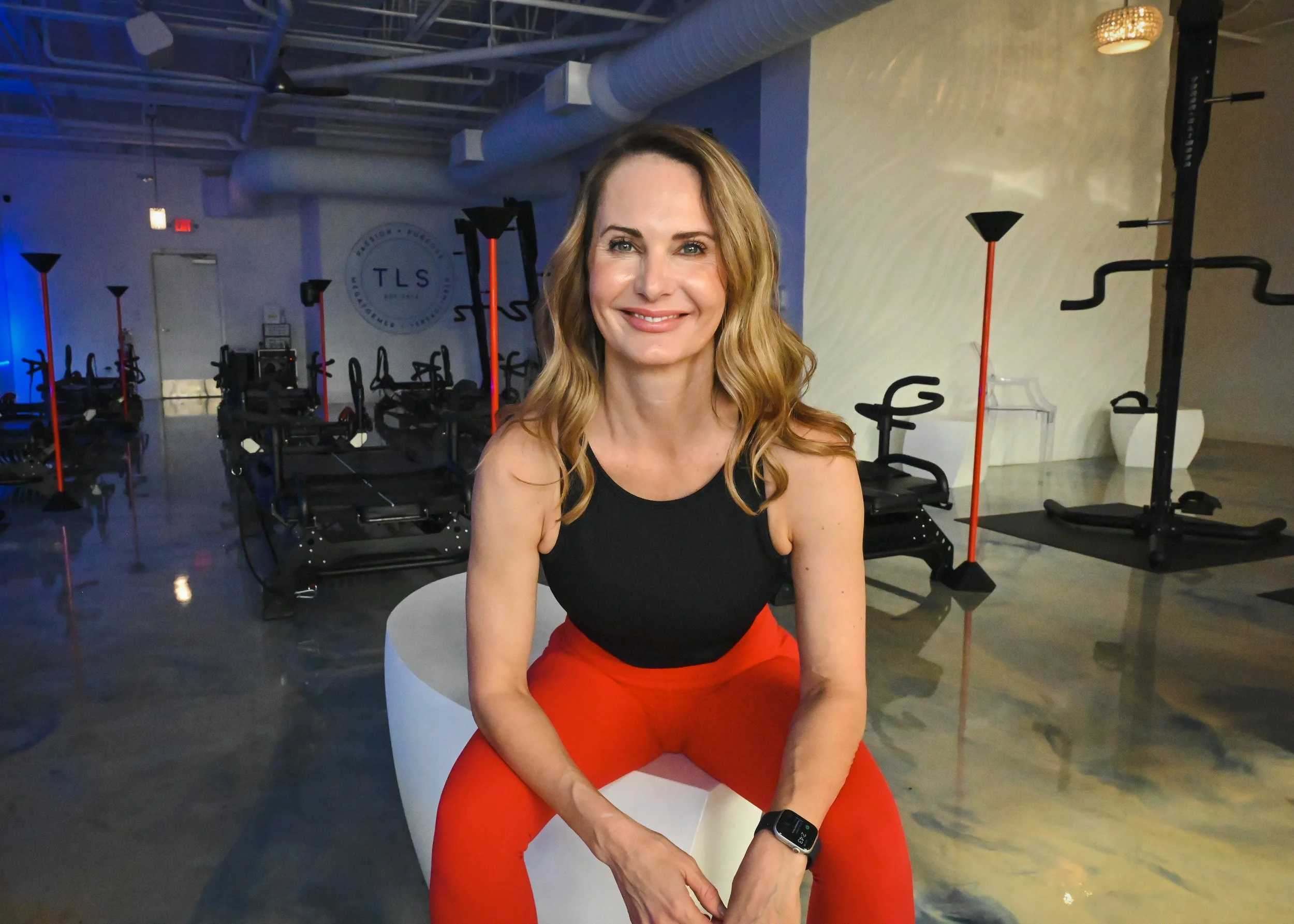 Woman sitting in a fitness studio with Megaformer equipment, wearing a black tank top and red leggings, smiling at the camera.