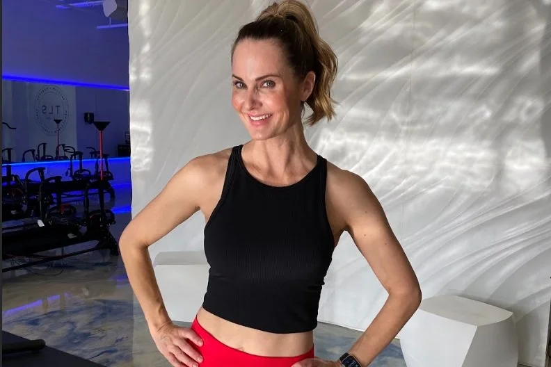 Woman in black crop top and red leggings posing in a gym environment with exercise equipment in the background.