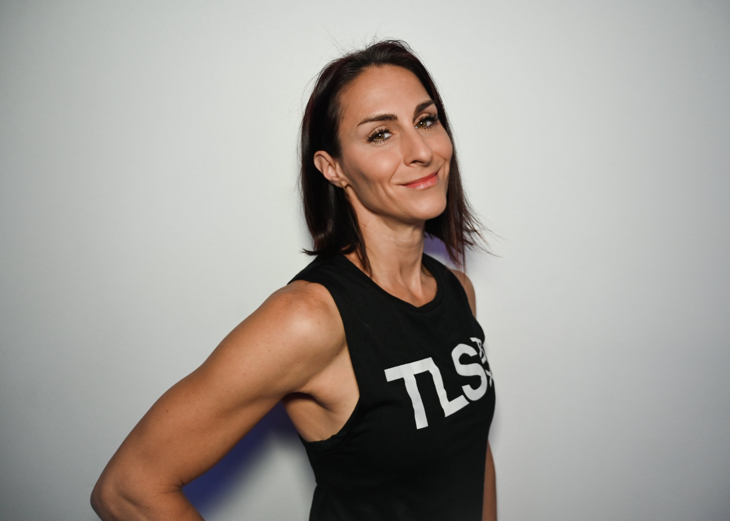 Woman wearing a black sleeveless top with the letters 'TLS' standing against a plain background.